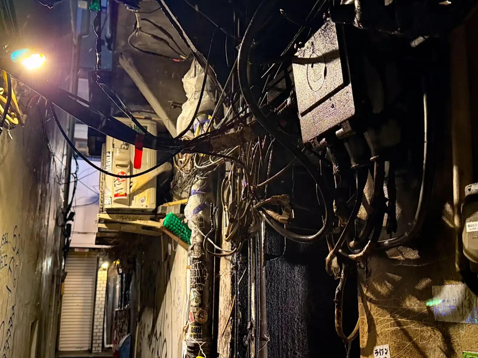 A thick tangle of black cables and an electrical box squeezed into the gap between two buildings.