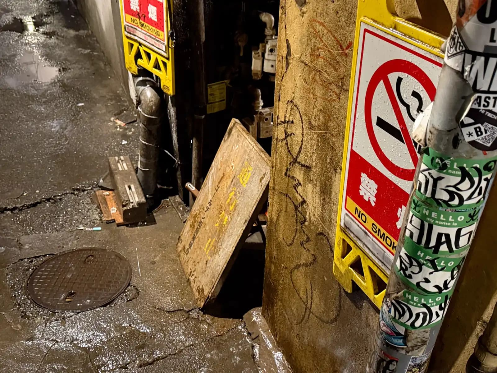 Wet, cracked pavement with a manhole cover, a couple of No Smoking signs and a pole plastered with name-tag stickers.