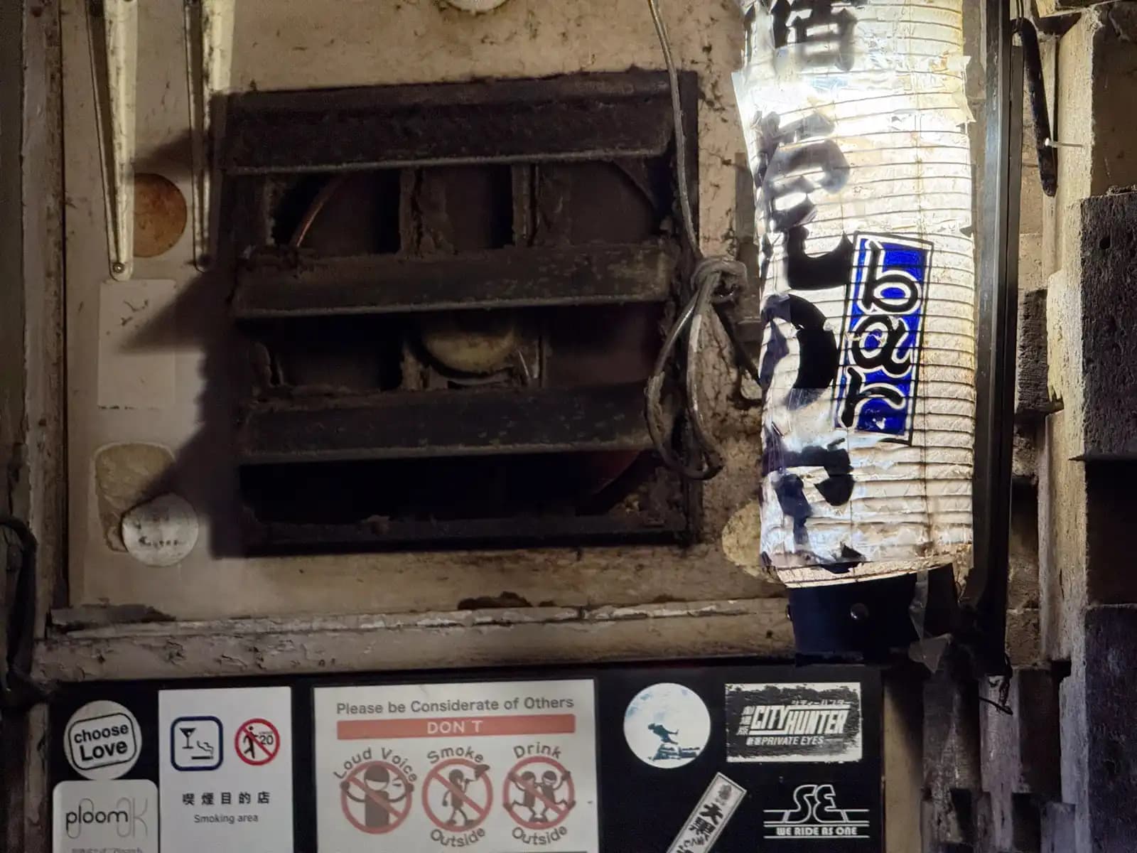 A paper lantern hangs next to a grimy old vent fan, above a row of stickers and house rules.