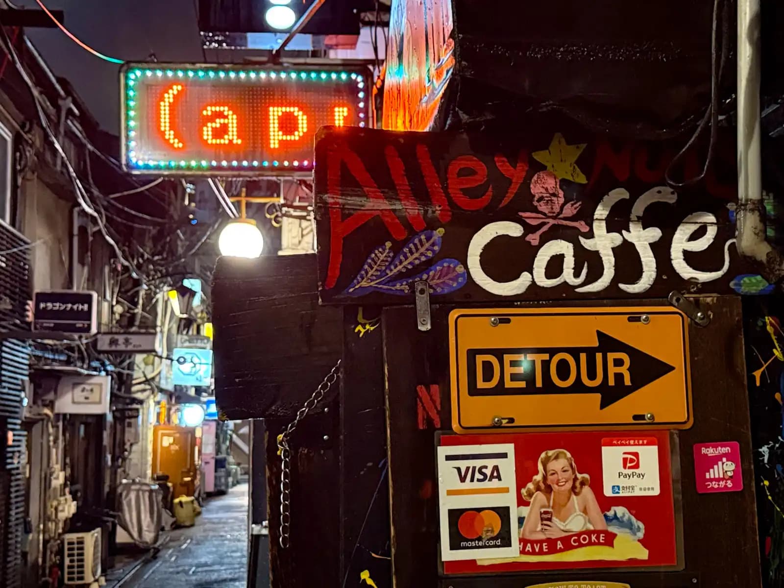 A hand-painted Alley Caffe sign with a yellow Detour arrow and an old Coke ad, next to a wet, empty alley.