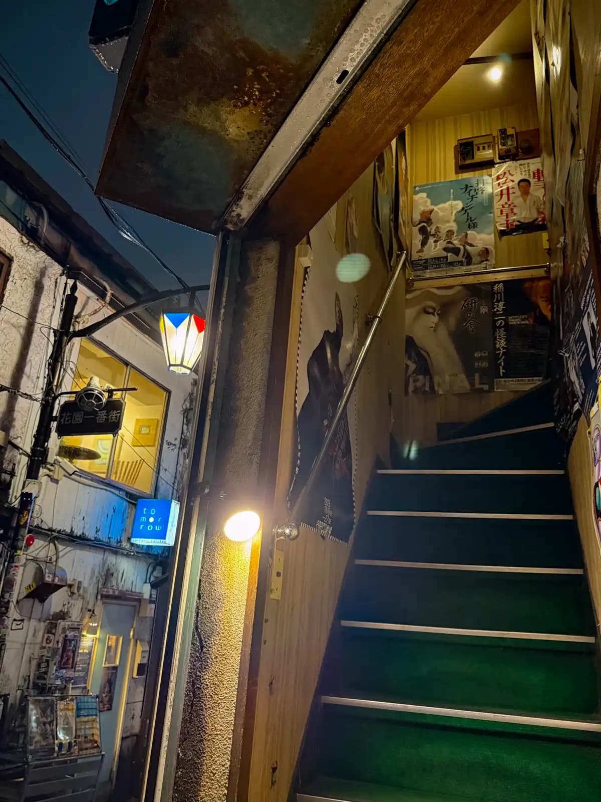 Green-carpeted stairs lined with old posters, with the Hanazono 1-bangai street sign and a lantern outside.
