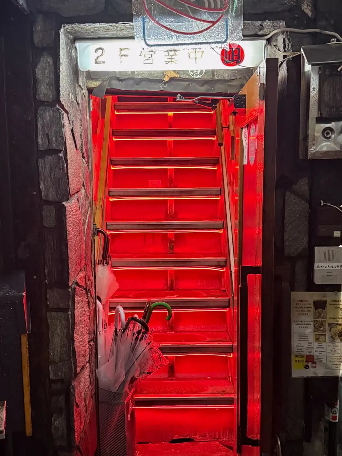 Steep stairs lit up bright red, with a sign saying the second-floor bar is open and wet umbrellas in a bin by the door.