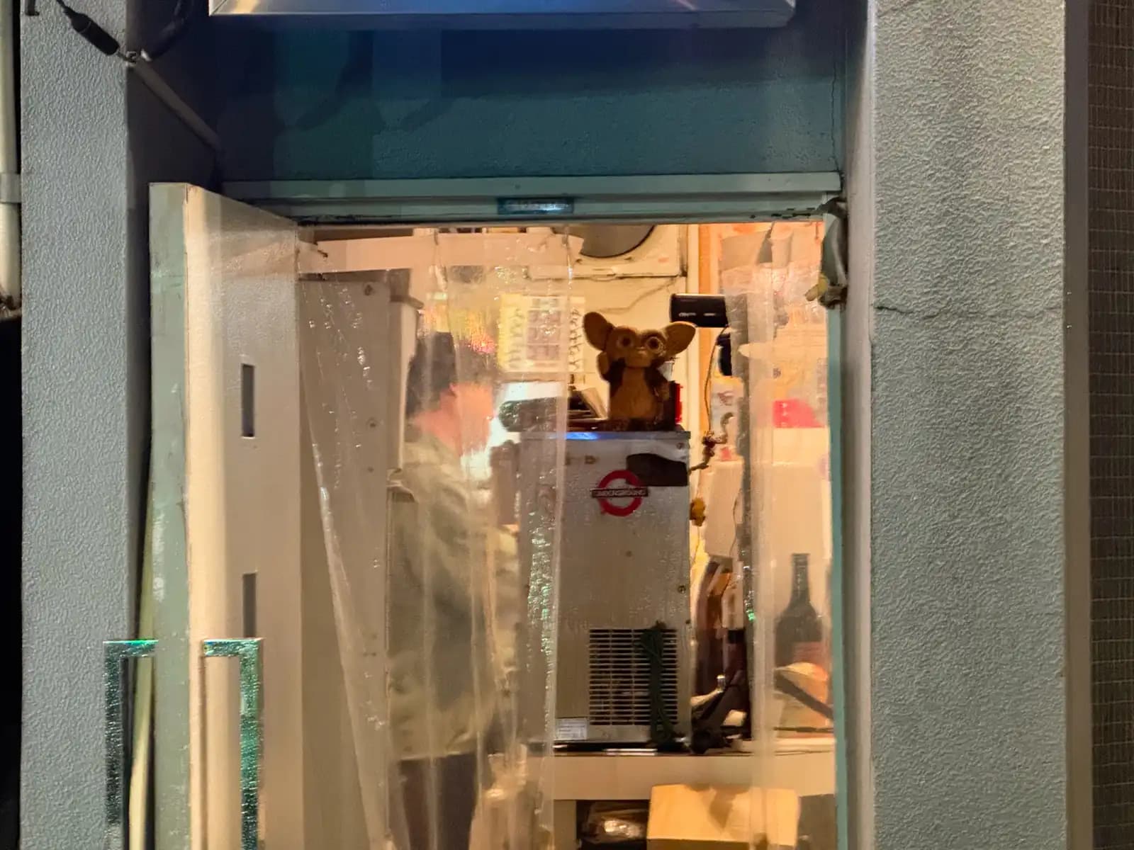 Looking through a plastic rain curtain into a tiny bar, where a Gremlin figure sits on top of the beer server.