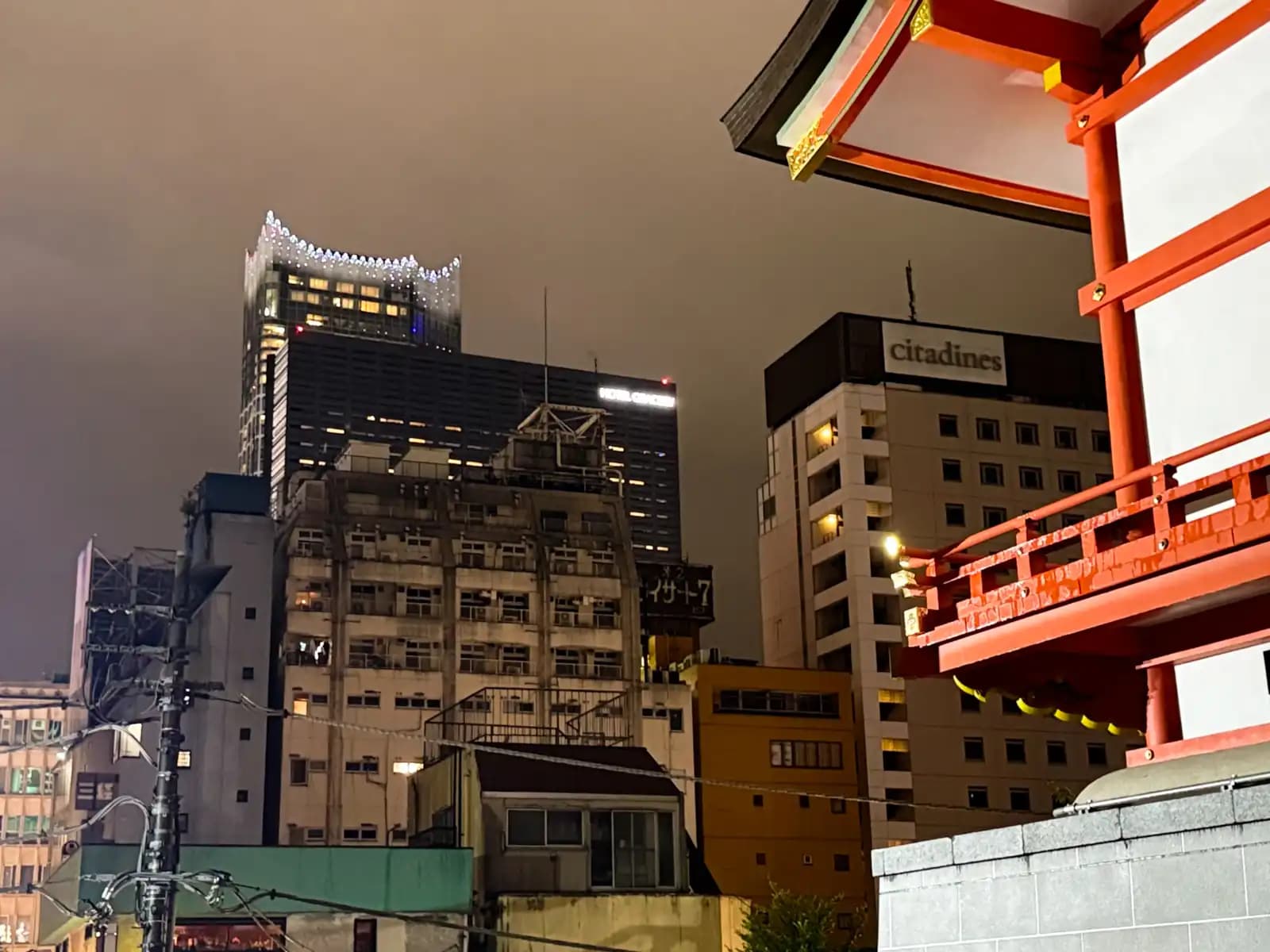 The vermilion eaves of Hanazono Shrine on the right, with the Tokyu Kabukicho Tower and the Citadines Central Shinjuku hotel lit up against the night sky behind them. Golden Gai sits down below, squeezed in between the shrine and the towers.