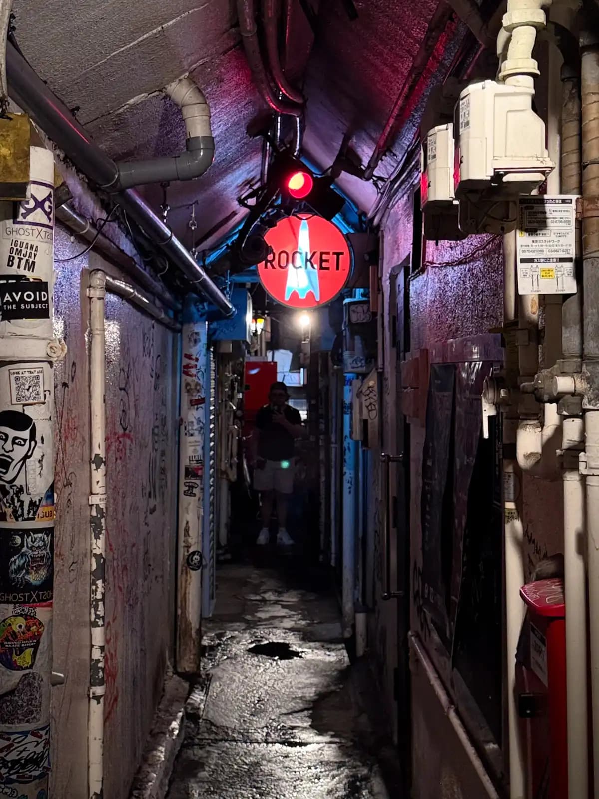 A cramped covered passage with a wet floor, pipes and stickers everywhere, and a red Rocket sign hanging from the ceiling.