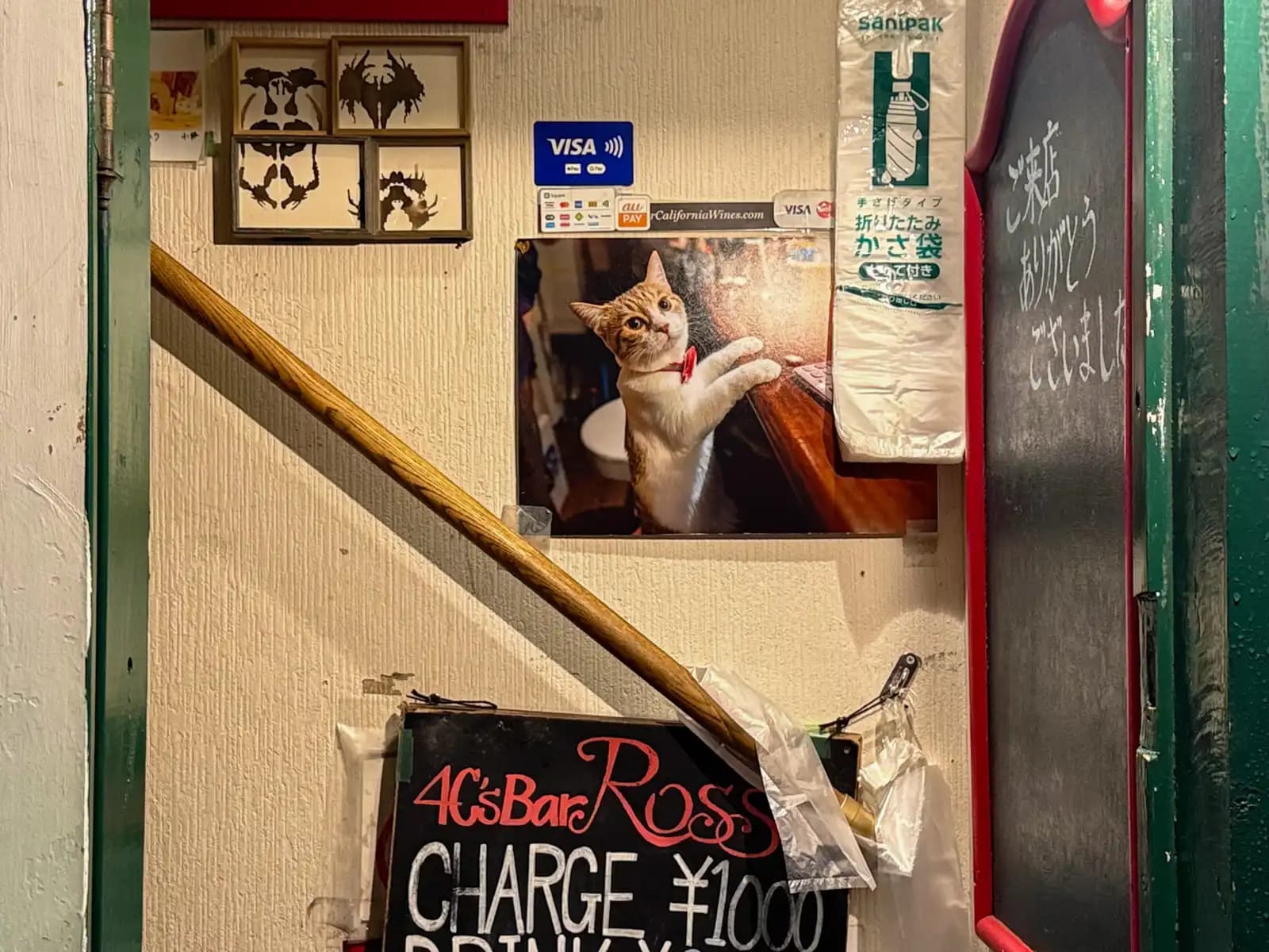 The entrance to 4C's Bar Ross: a ¥1,000 charge sign, a photo of a cat at the bar and framed inkblot pictures by the stairs.