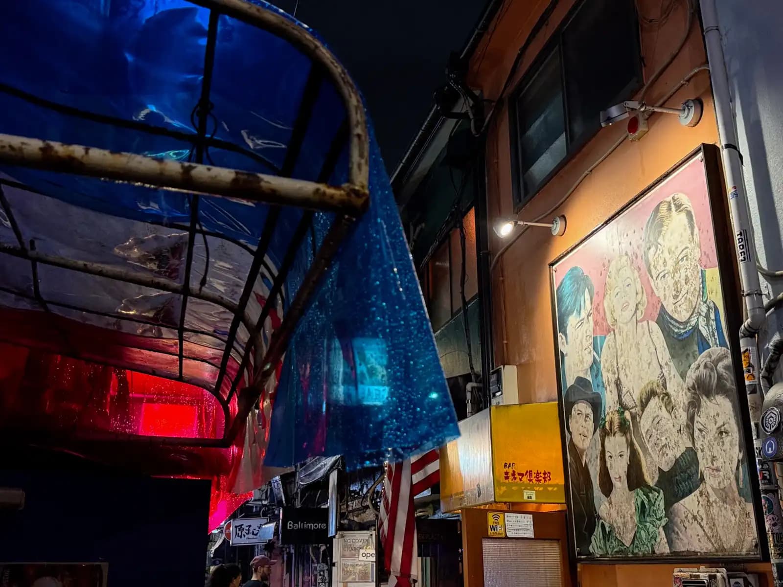 A big painting of old movie stars on the side of a bar, next to a blue plastic awning covered in raindrops.
