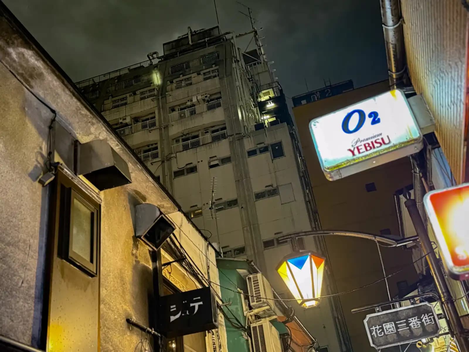Looking up at an O2 sign with a Yebisu beer logo, a colorful street lamp and the Hanazono 3-bangai sign.