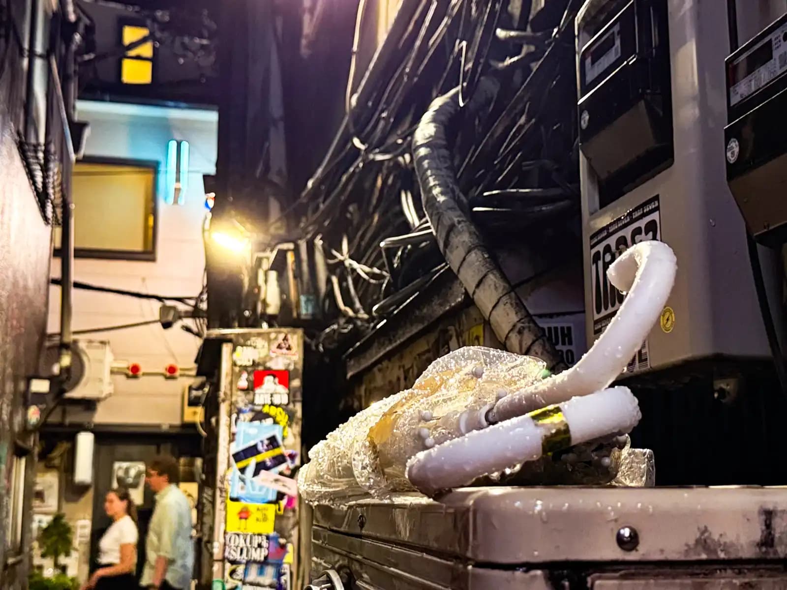 A couple of wet plastic umbrellas left on a metal box under the electric meters, with people walking by in the back.
