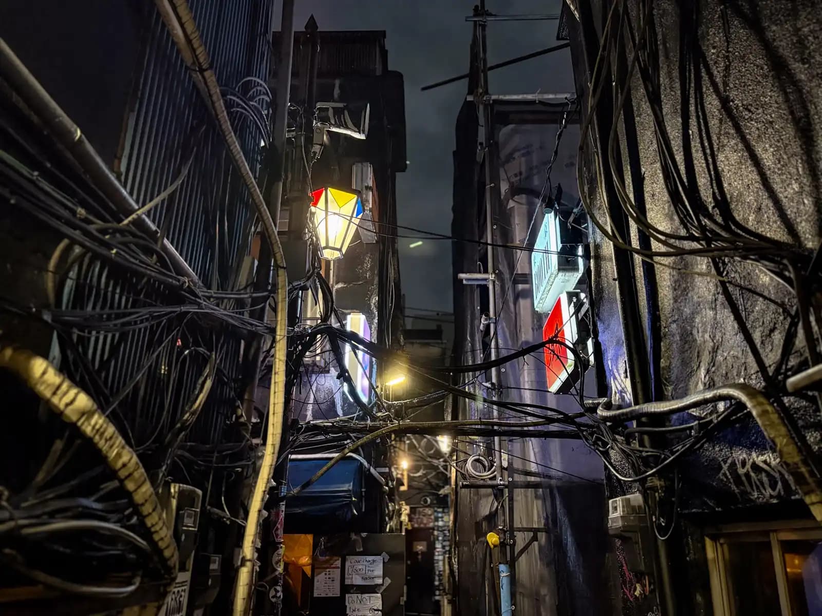Looking up a dark, narrow gap full of loose cables, with a colorful lamp and a few bar signs lit up.