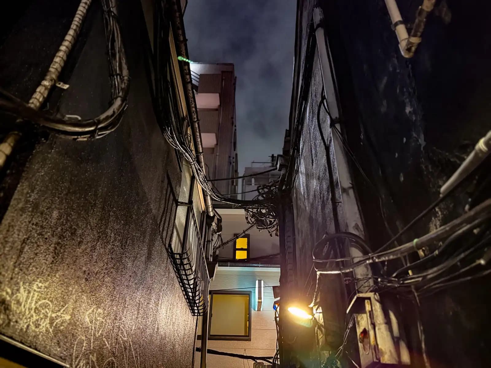 Looking straight up between two dark, damp walls covered in wires, toward a cloudy night sky.
