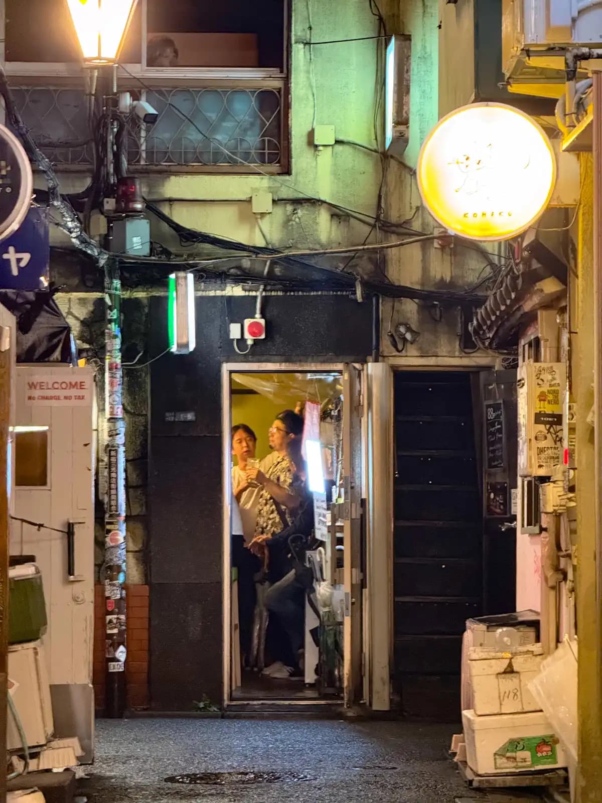 People having drinks inside a tiny bar with the door wide open, next to a "Welcome, no charge, no tax" sign.