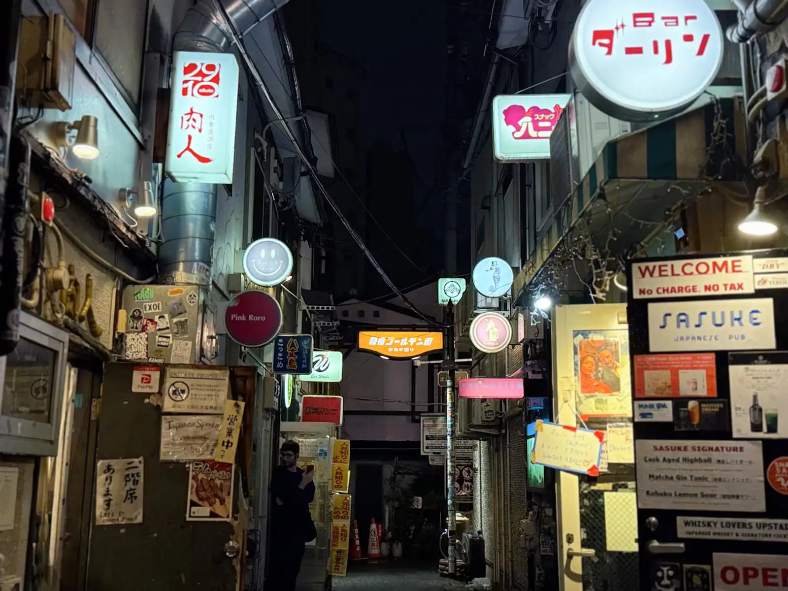 An alley full of lit signs, including Bar Darling, Nikujin, Pink Roro and Sasuke, whose board says no charge, no tax.