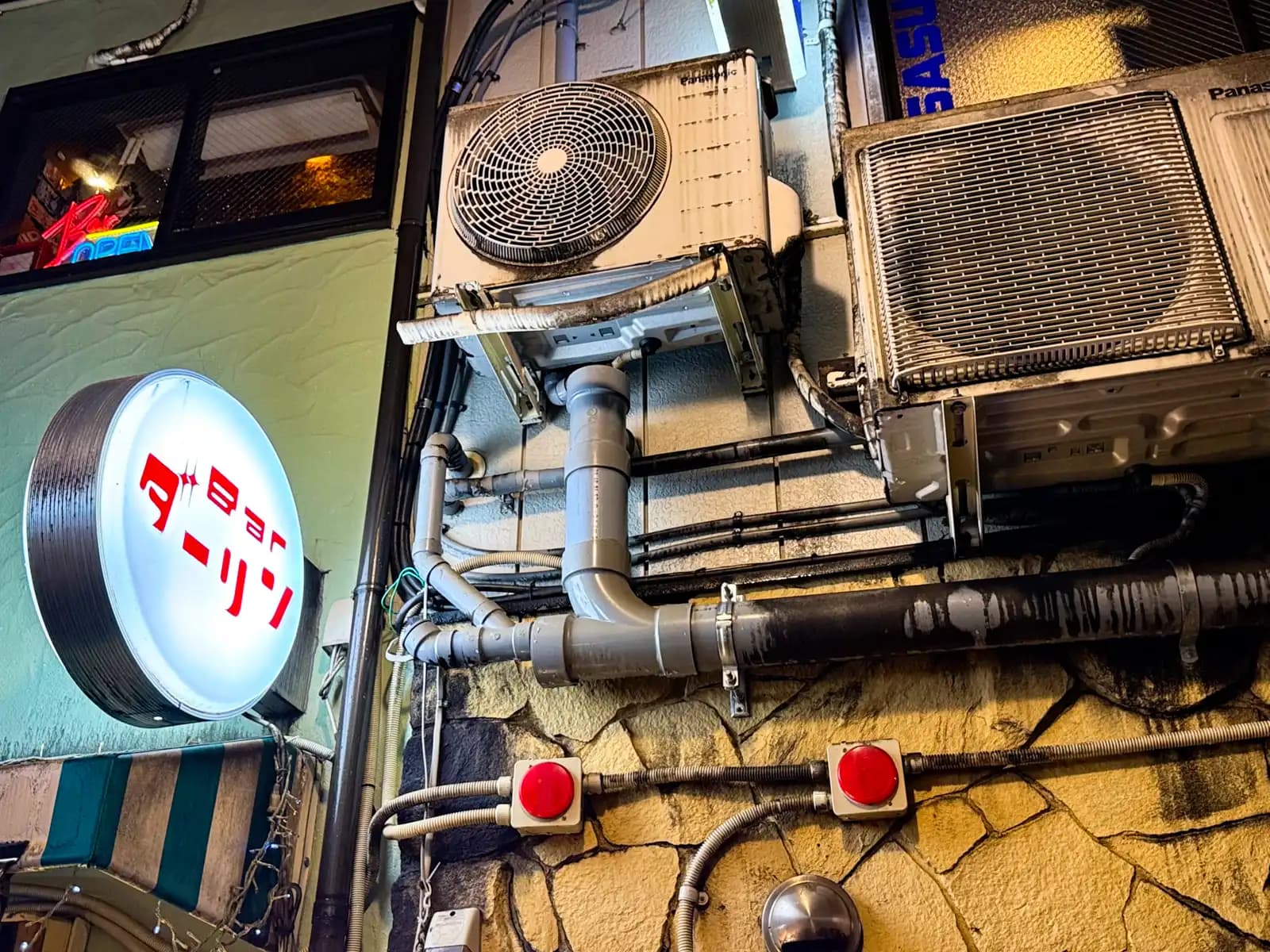 The round sign for Bar Darling next to a couple of grimy air conditioner units and a tangle of pipes on a stone wall.