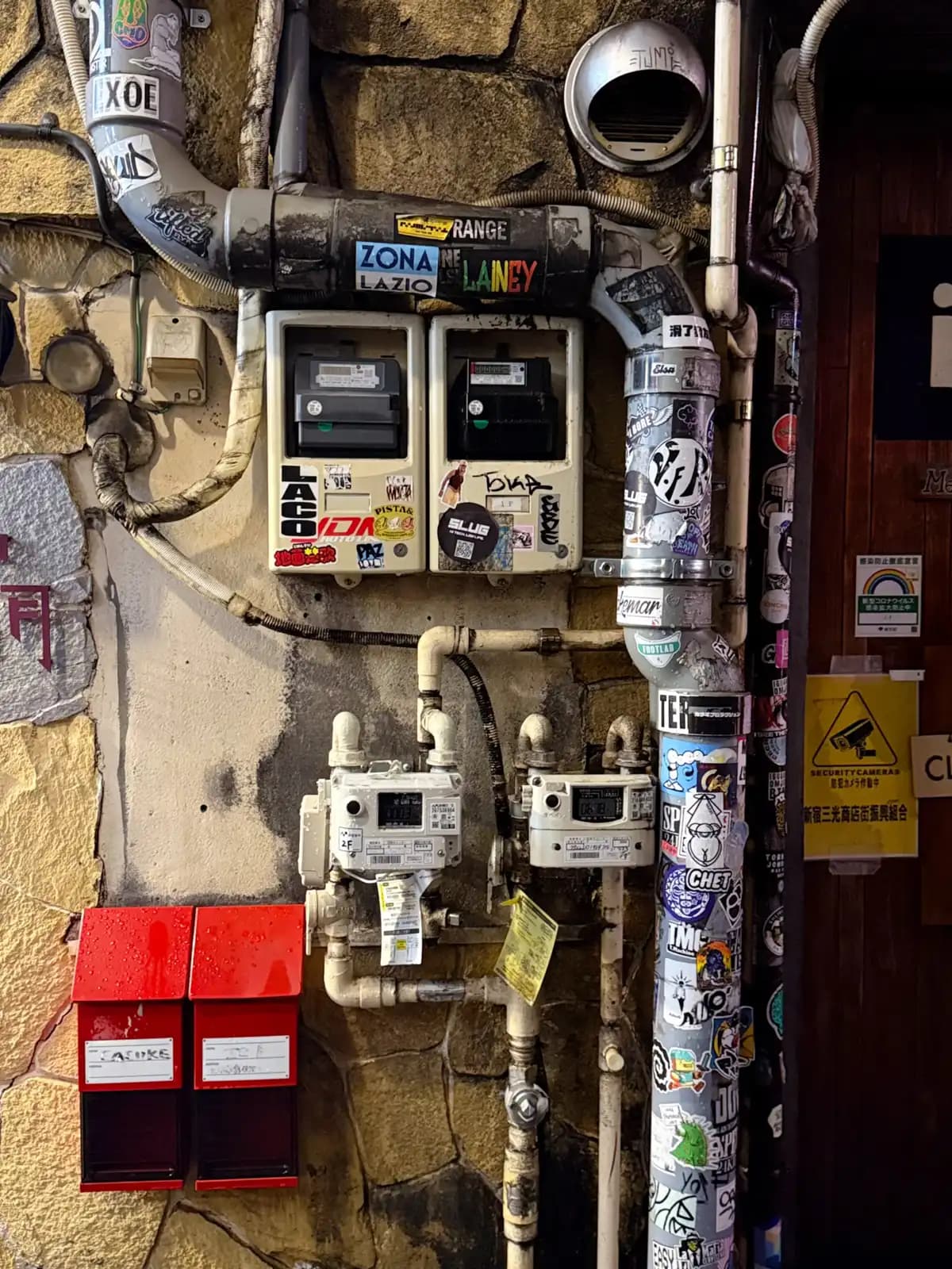 Utility meters and pipes plastered with stickers, above two red mailboxes on a stone wall.