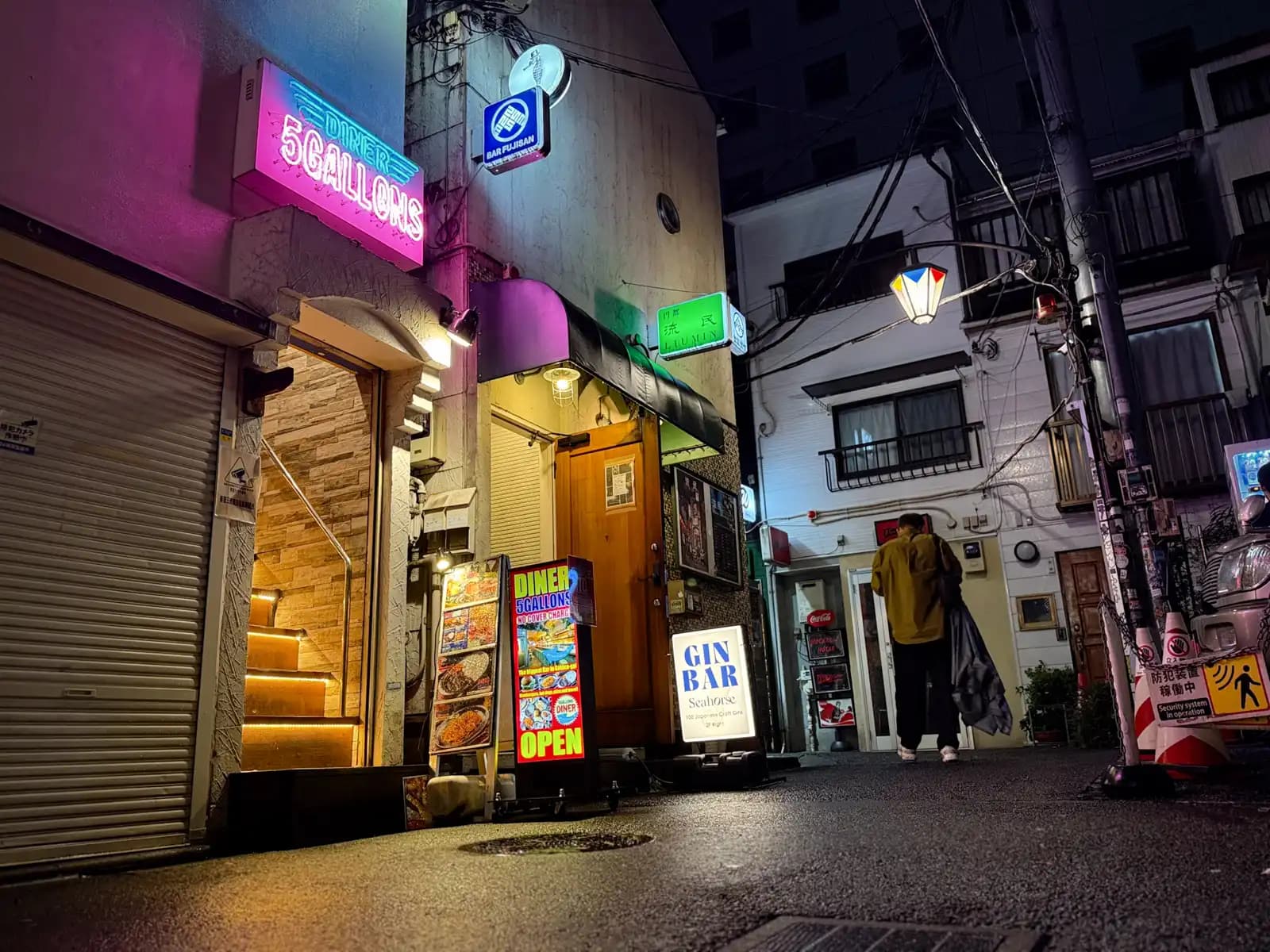 The pink neon sign of 5 Gallons Diner, with Bar Fujisan and Gin Bar Seahorse next door, and a man walking by.