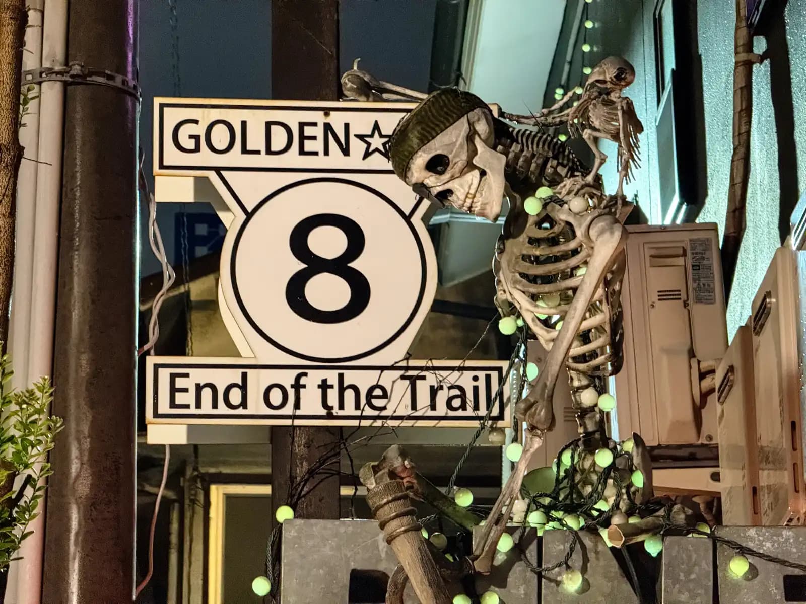 A plastic skeleton in a knit cap leans on a "Golden Gai 8 End of the Trail" road sign, strung with green lights.