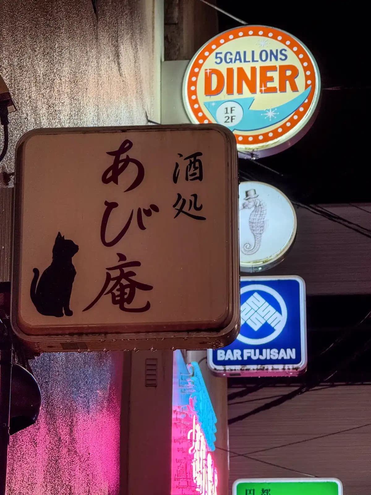 A sign for the bar Abian with a black cat on it, stacked with signs for 5 Gallons Diner, a seahorse and Bar Fujisan.
