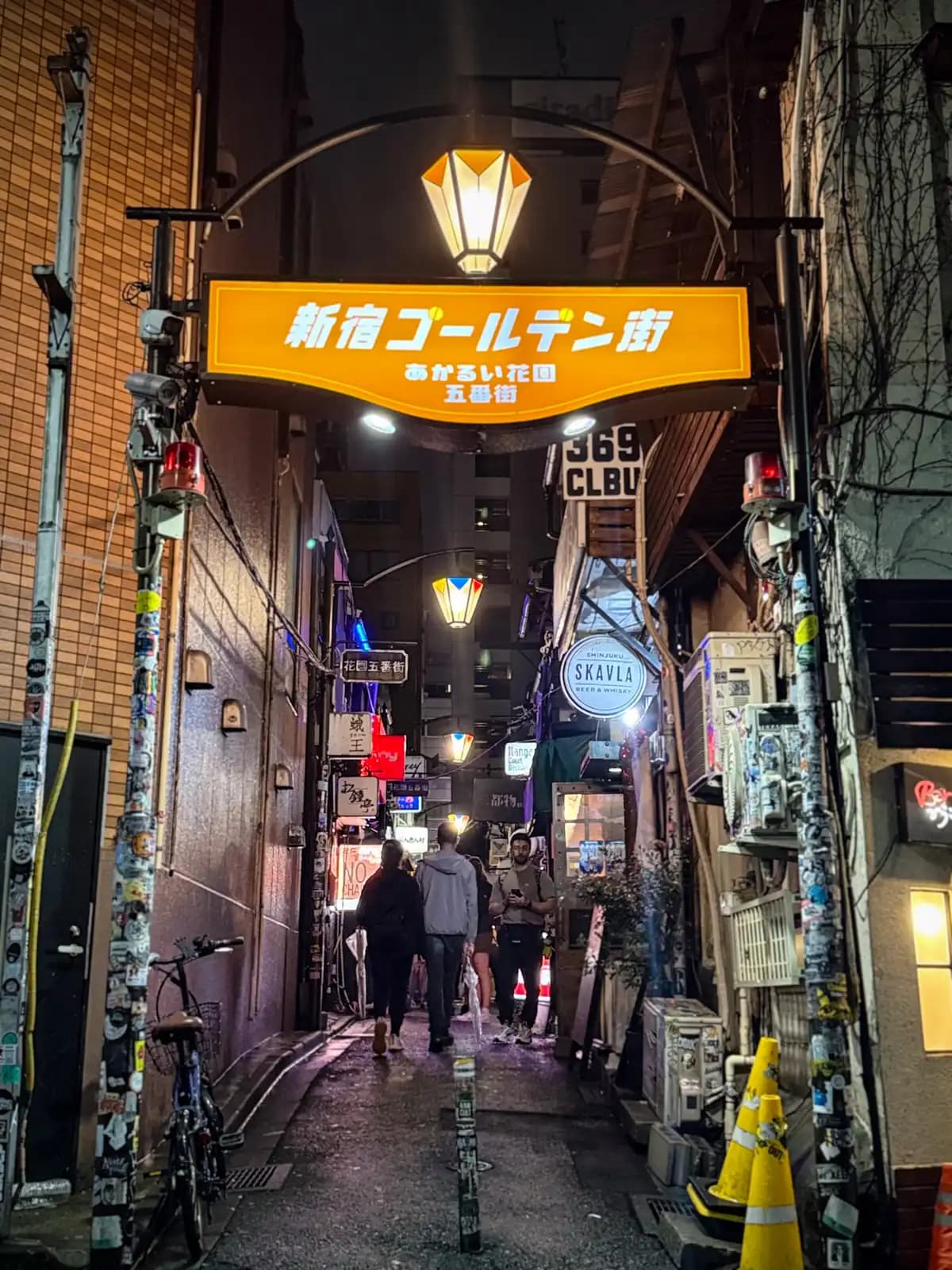 The orange Shinjuku Golden Gai sign over the entrance to Hanazono Gobangai, with people heading into the alley past SKAVLA.