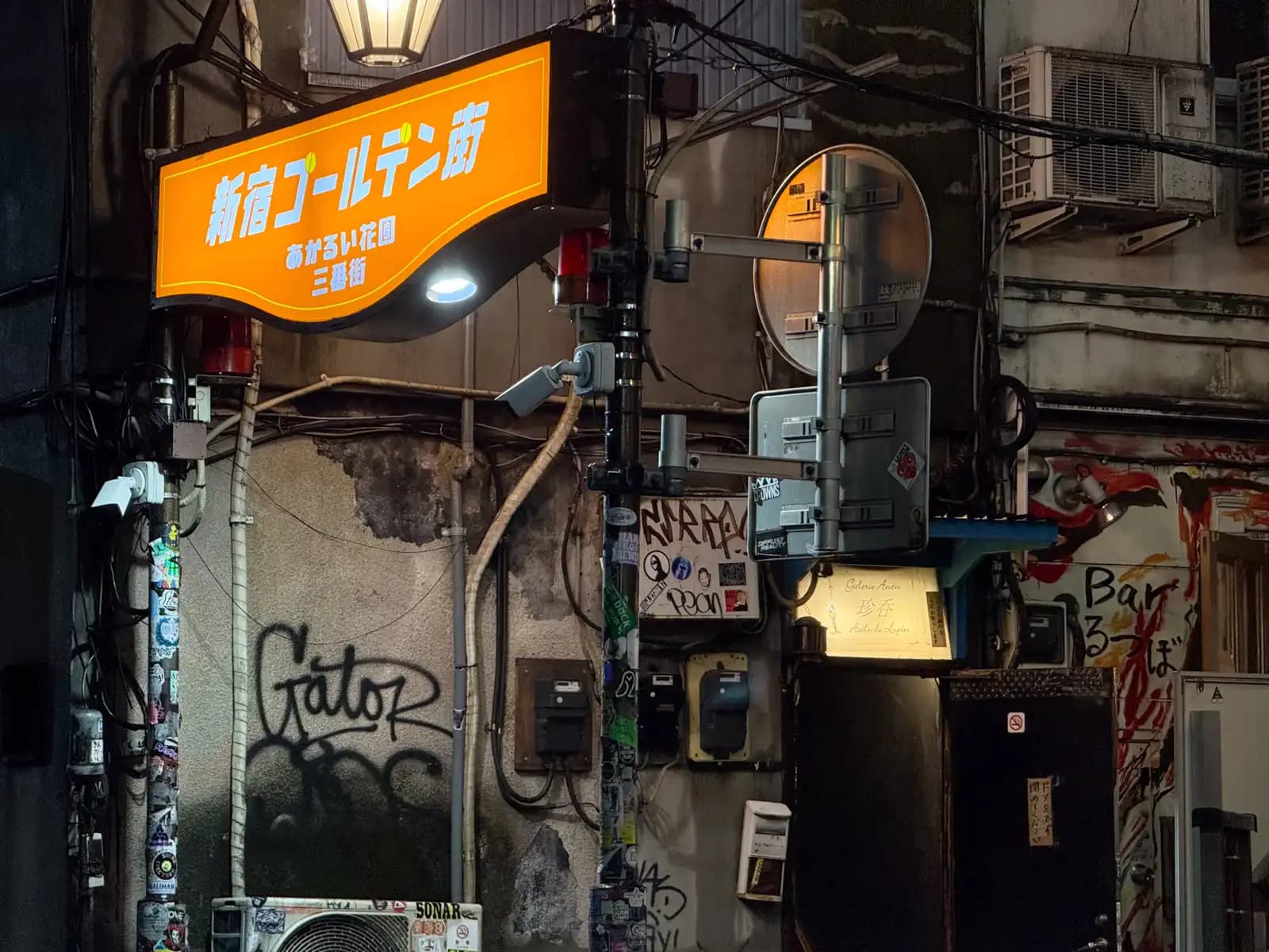The orange sign for Hanazono Sanbangai over a beat-up wall with "Gator" graffiti, meters, cameras and a painted bar sign on the right.