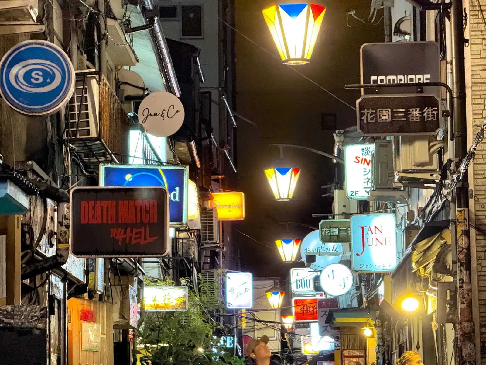 Looking down Hanazono Sanbangai at a pile of lit-up bar signs, including Death Match in Hell and Jan June, under colorful street lamps.