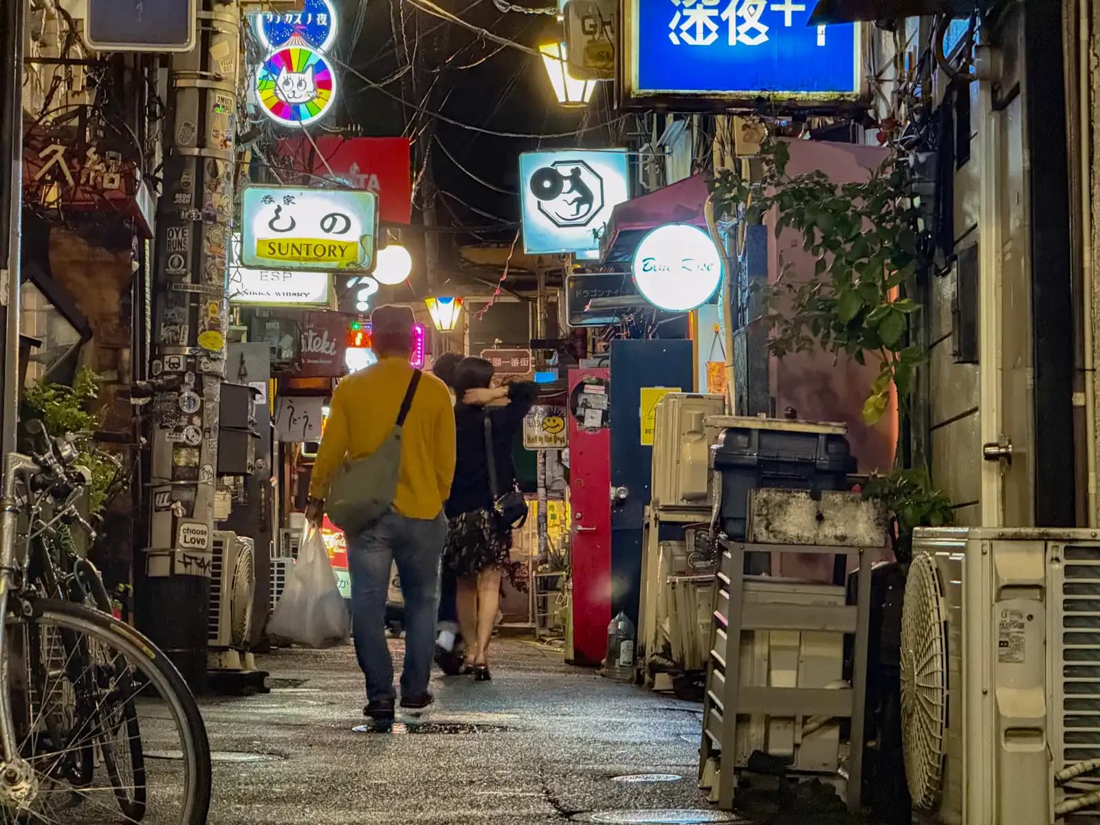 People walk down a wet alley past bicycles and air conditioners, with signs for Shino and Shinya+1 lit up overhead.