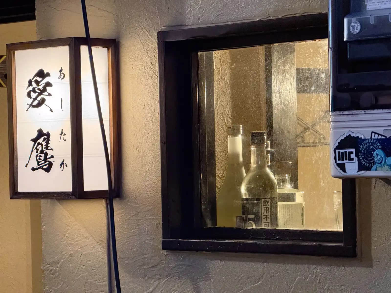 A paper lantern sign for the bar Ashitaka next to a small window with a few bottles behind the glass.