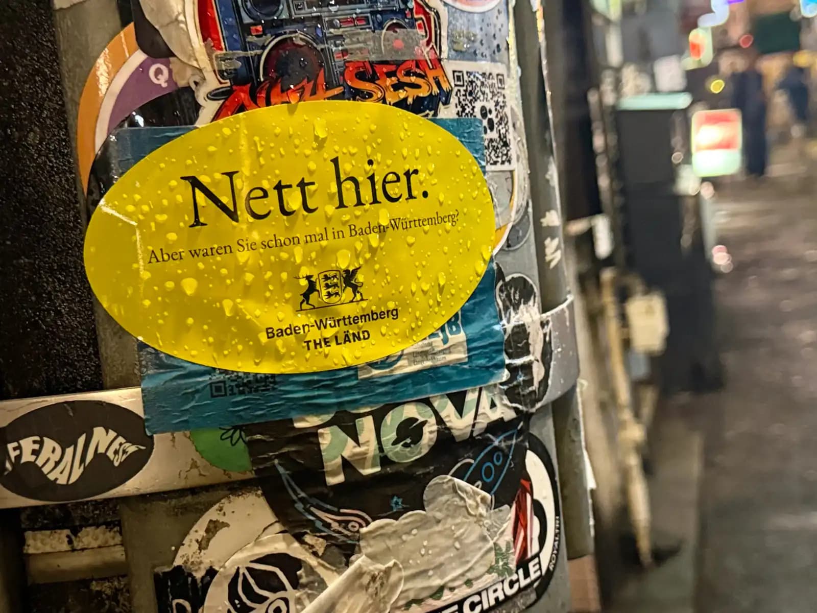 A wet yellow sticker on a pole says "Nett hier. Aber waren Sie schon mal in Baden-Württemberg?" ("Nice here. But have you ever been to Baden-Württemberg?"), a German tourism ad among the other stickers.