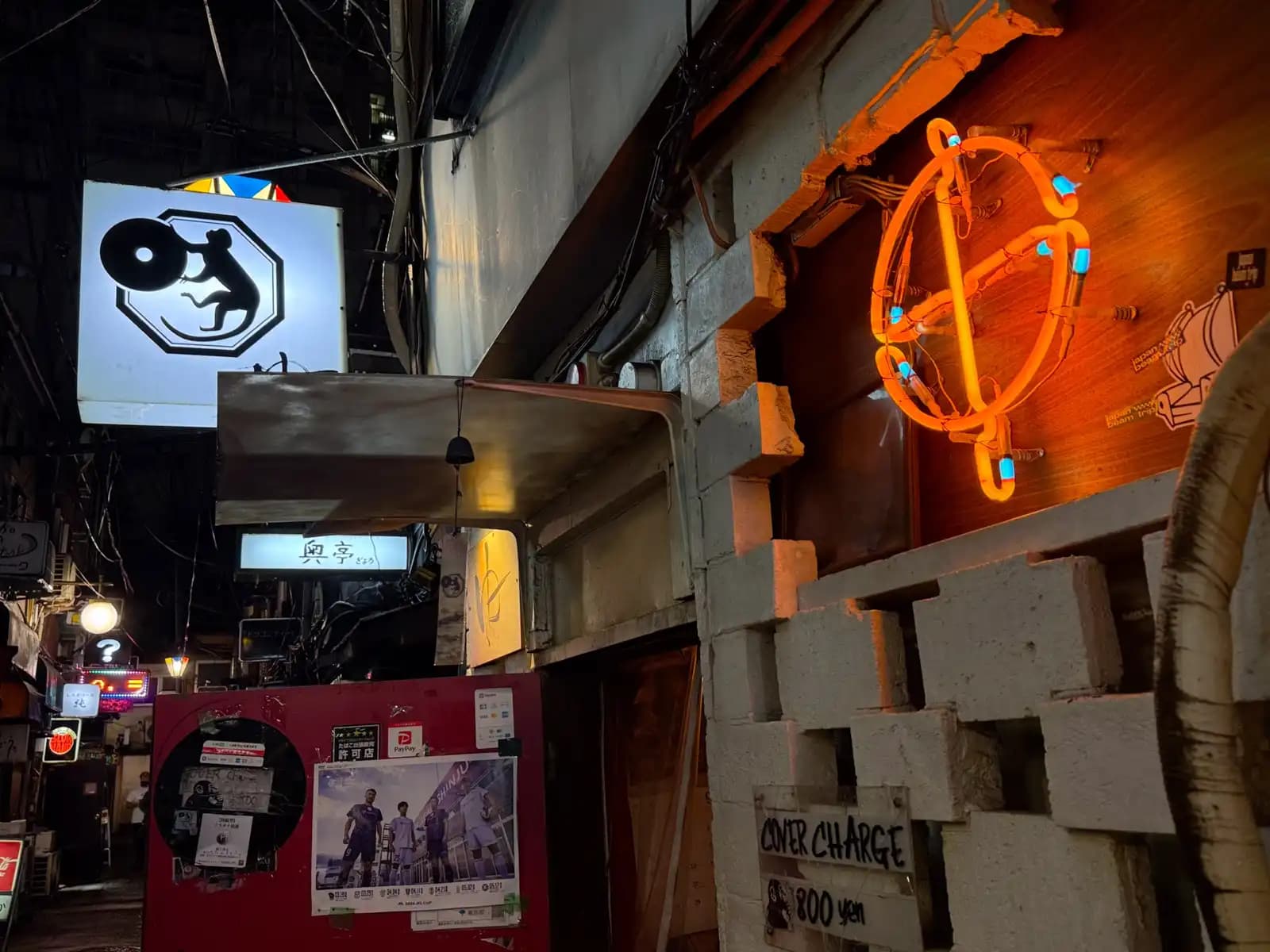 An orange neon sign on a cinder block wall with an 800 yen cover charge, and a monkey-with-a-record sign hanging nearby.