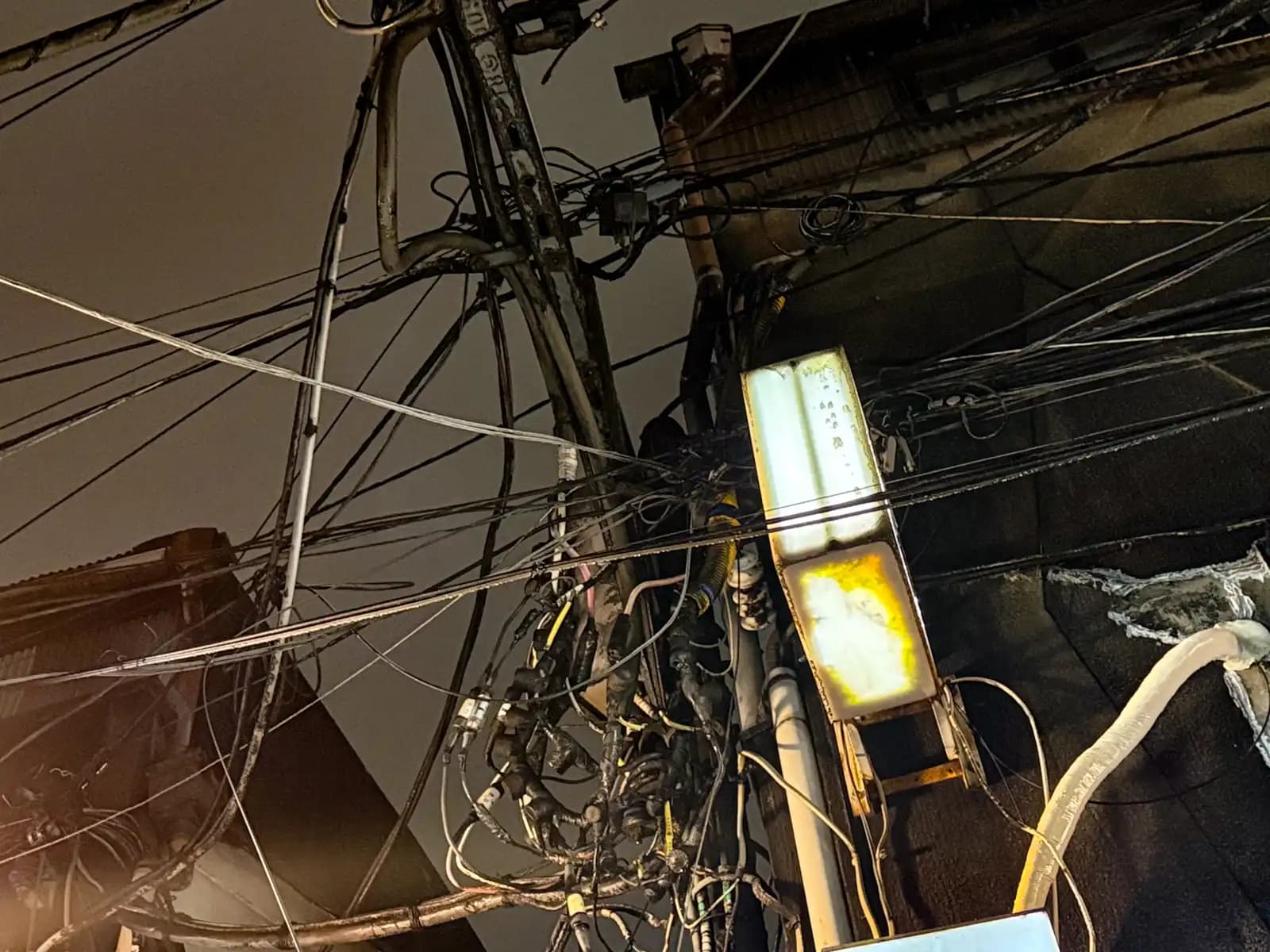 A mess of power lines and cables wrapped around a utility pole, with an old light box stuck to the side.