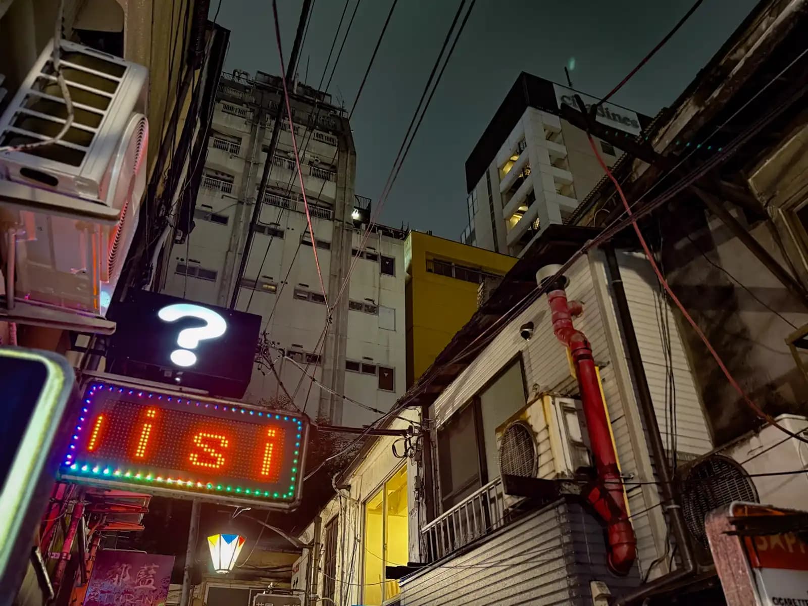 A glowing question mark sign and an LED sign in the alley, with taller office buildings rising behind the old bars.