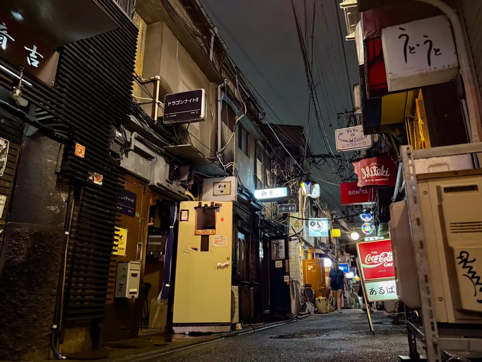 Low view down a wet, quiet alley with signs for Dragon Night, Bonita and a Coca-Cola sign, and one woman walking our way.