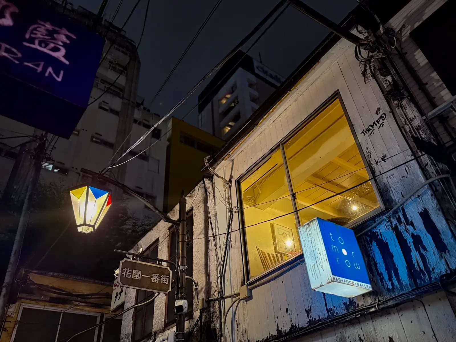 The blue sign for tomorrow, a bar, food and gallery spot, next to a lit-up window, a street lamp and the Hanazono Ichibangai sign.