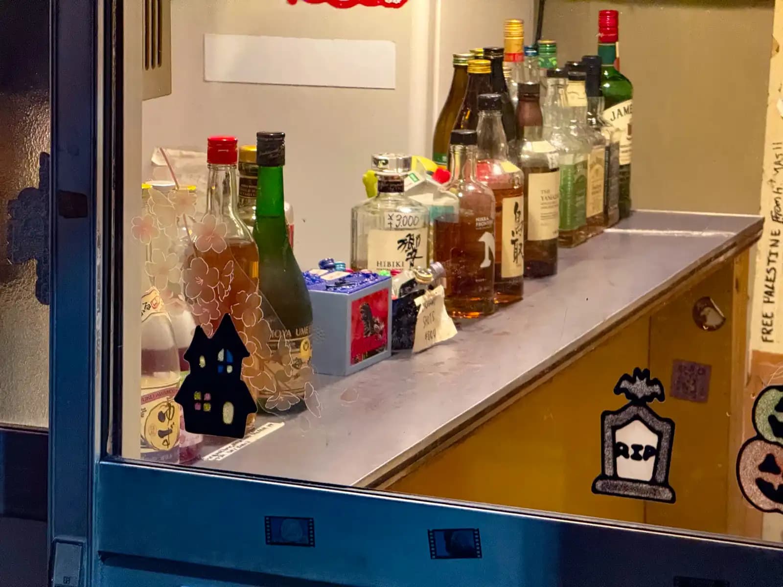 Bottles lined up on a bar counter seen through the window, including Hibiki priced at 3,000 yen, with Halloween stickers on the glass.