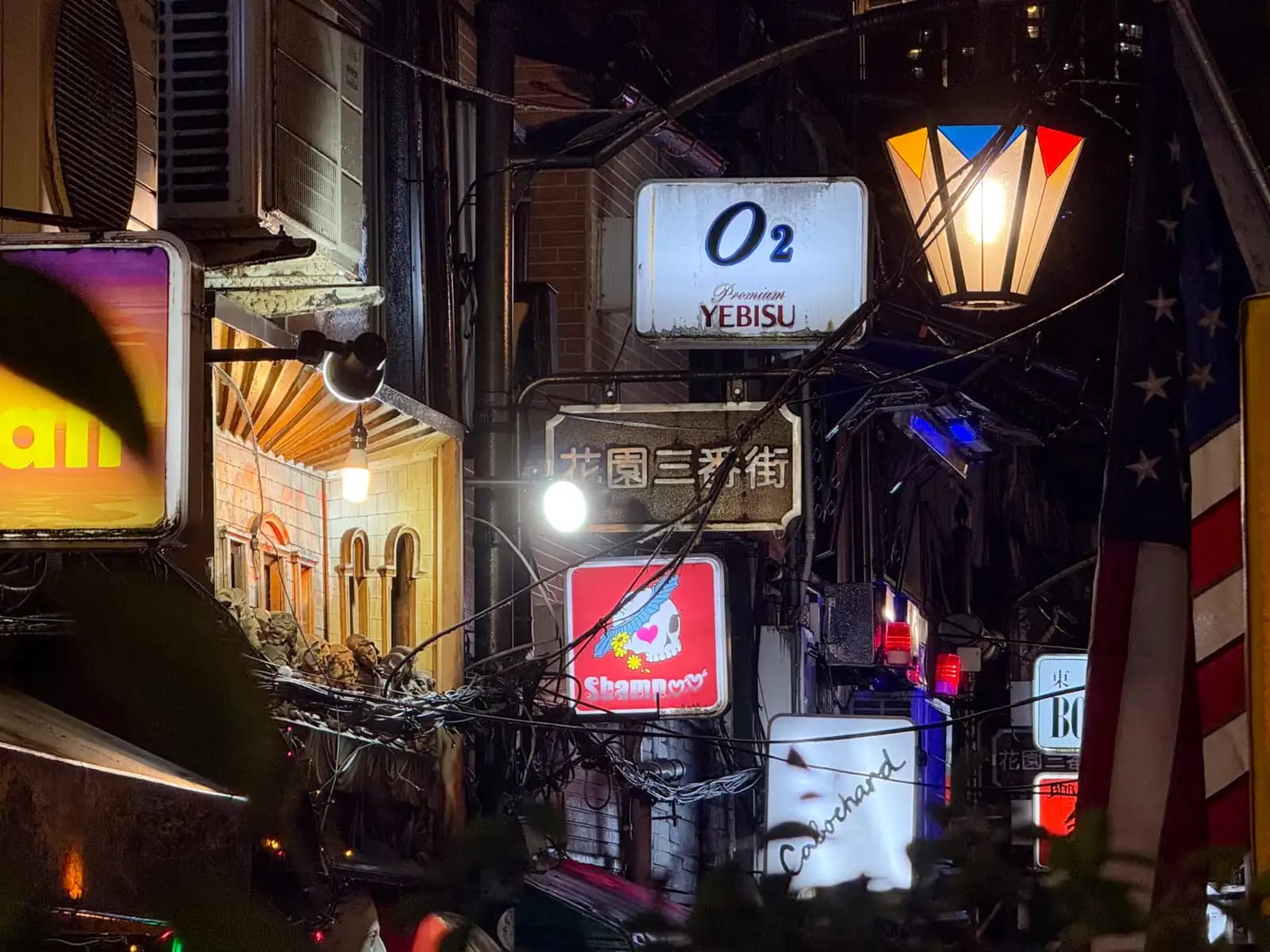 Stacked signs in Hanazono Sanbangai, including O2 with a Yebisu logo and a red skull sign, with an American flag hanging on the right.