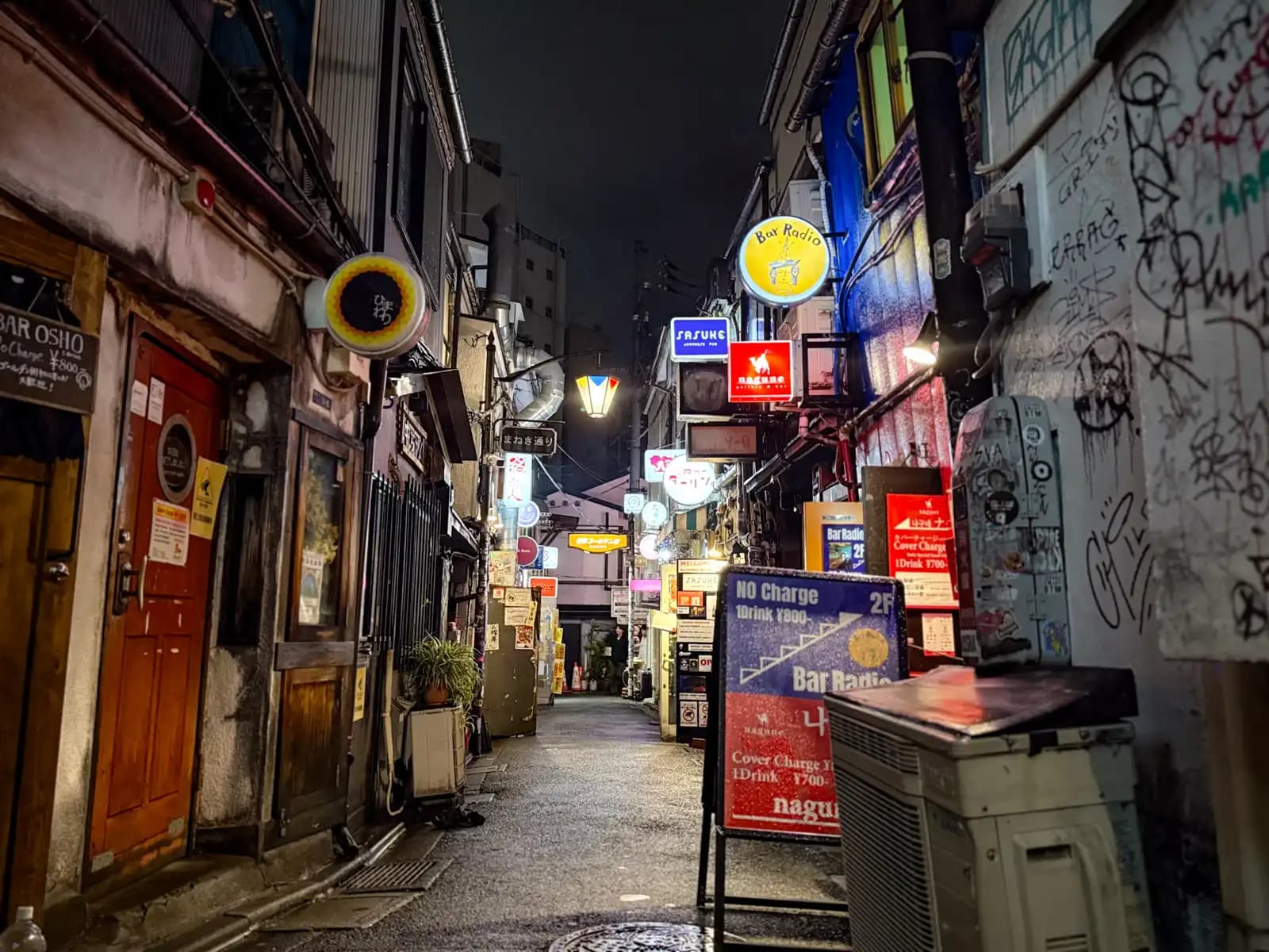 A long view down the alley at night, with Bar Osho on the left and Bar Radio, Sasuke and Nagune on the right past a graffiti-covered wall.