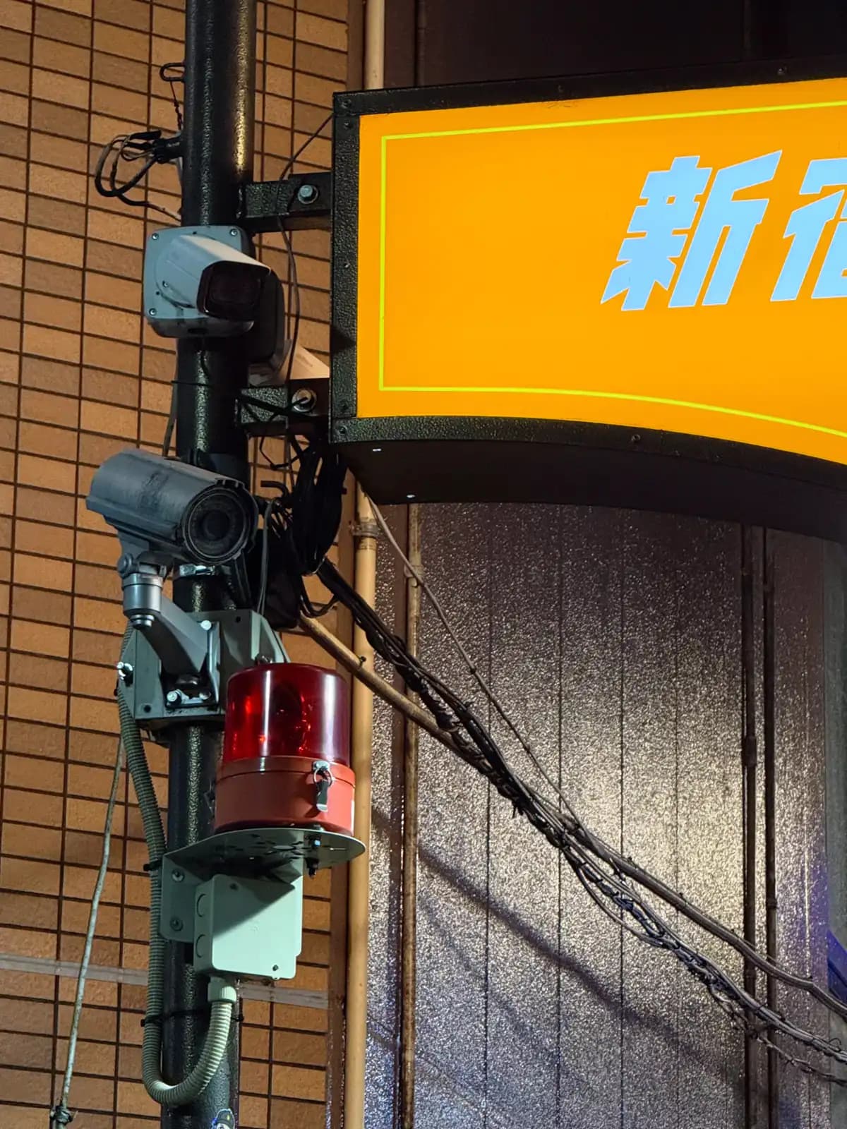 Security cameras and a red warning light on a pole next to the orange Shinjuku Golden Gai sign.