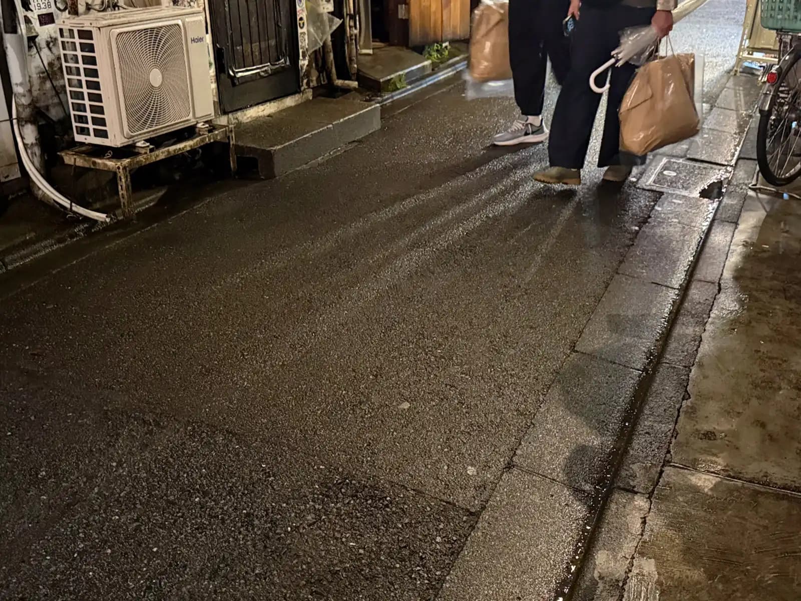 Wet pavement after the rain, with people walking out of frame carrying paper bags and an umbrella.