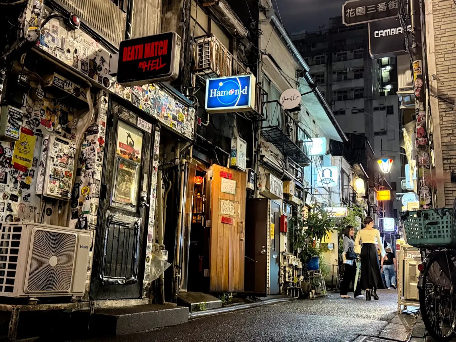 A sticker-covered bar front under the Death Match in Hell sign, with Hamond next door and two women walking up Hanazono Sanbangai.
