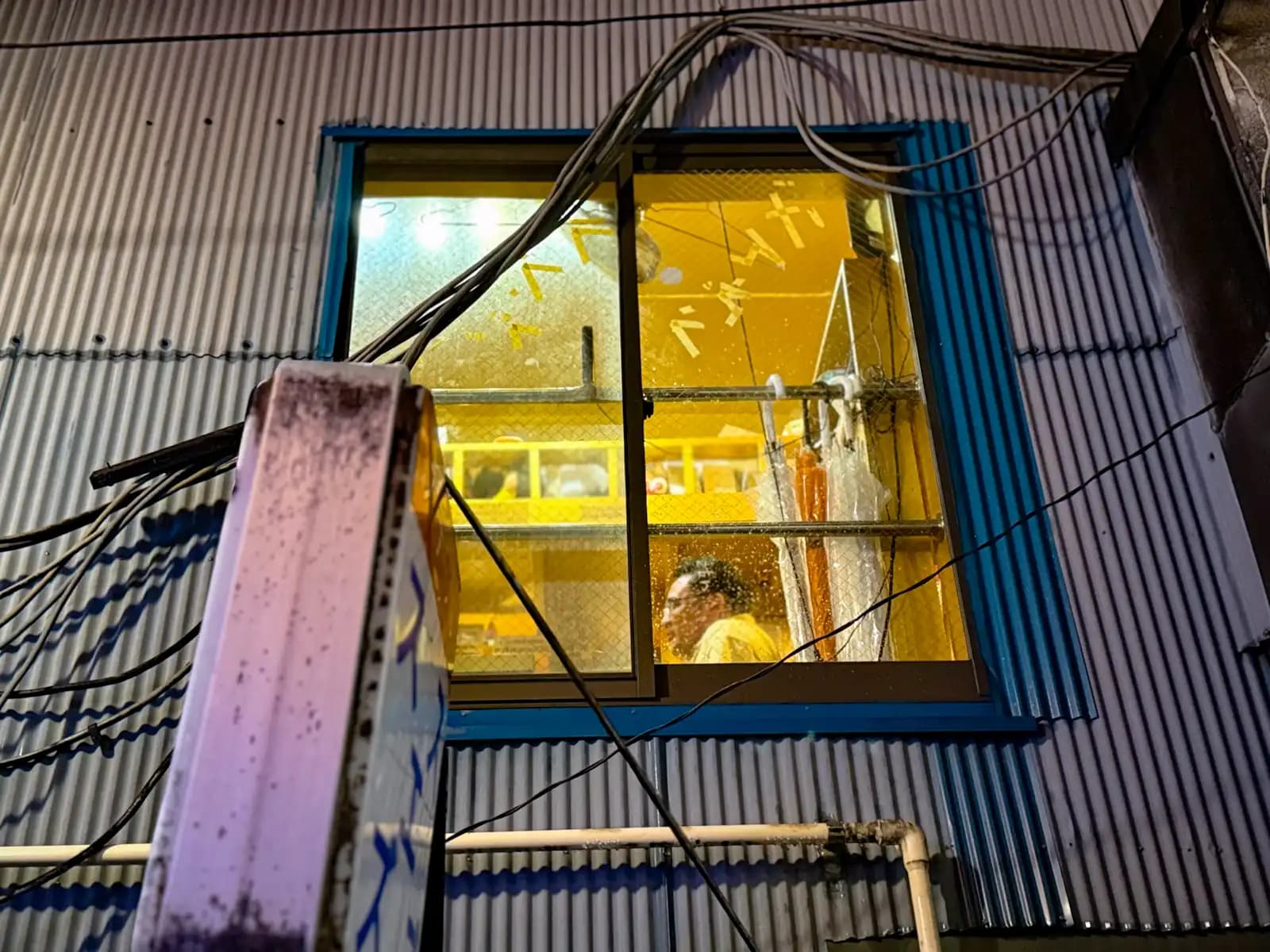 Looking up at a bright yellow second-floor window in a corrugated metal wall, with a man sitting inside and cables hanging in front.