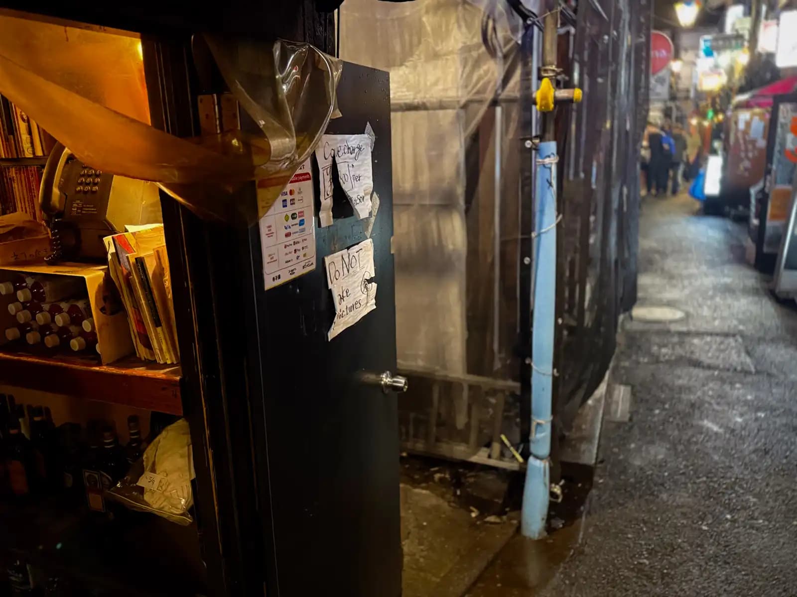 Handwritten notes on a bar door about the cover charge and "Do NOT take pictures," with the wet alley and people further down.