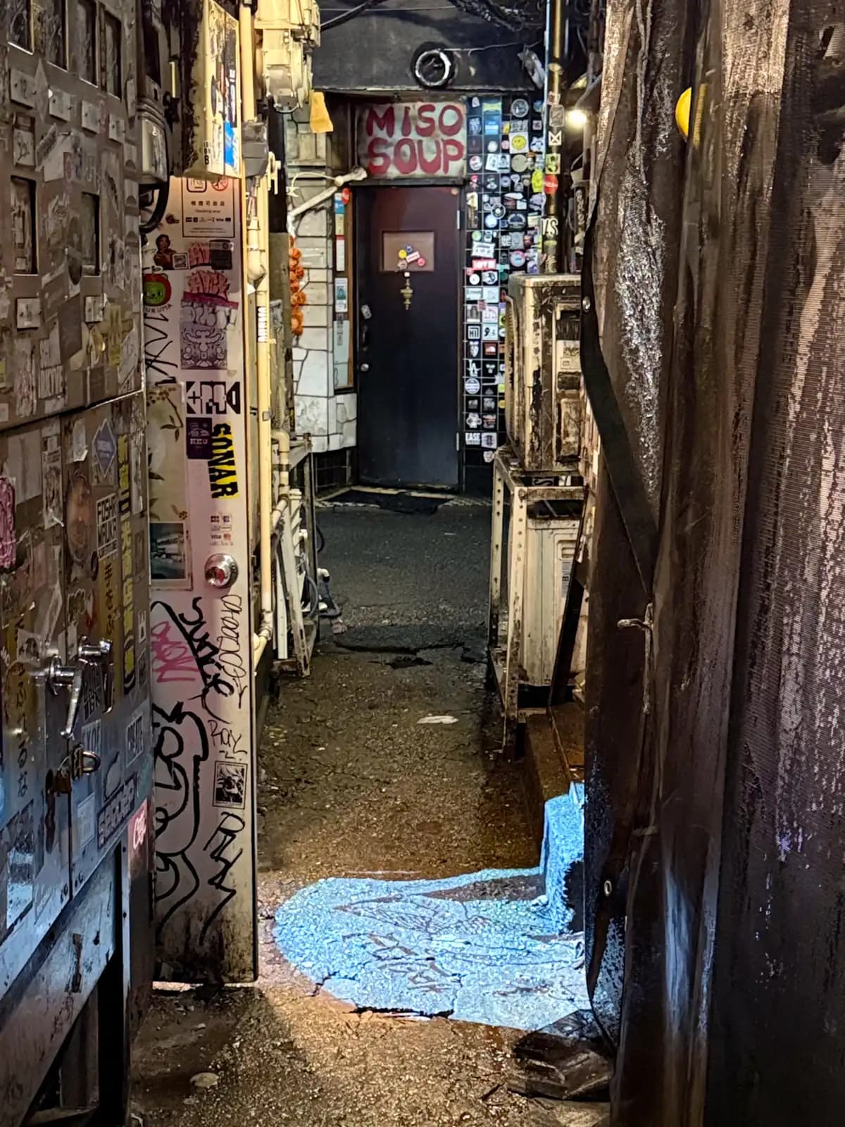 A cramped passage between buildings leading to a sticker-covered door under a hand-painted "MISO SOUP" sign.