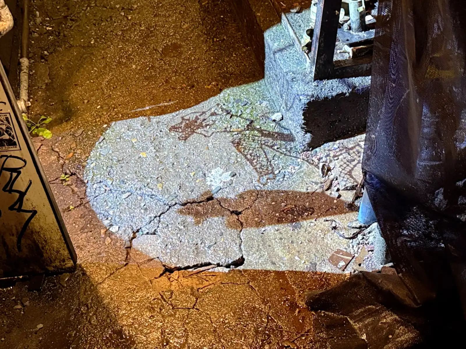 A light projects a slowly rotating pattern onto the cracked concrete ground, next to a black tarp and a graffiti-covered box.