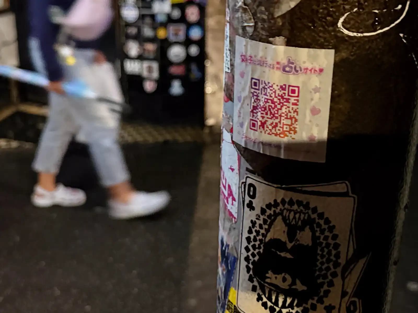 Stickers on a pole, including a QR code for a fortune-telling site and a queen of clubs with a big toothy mouth, as someone walks by with an umbrella.
