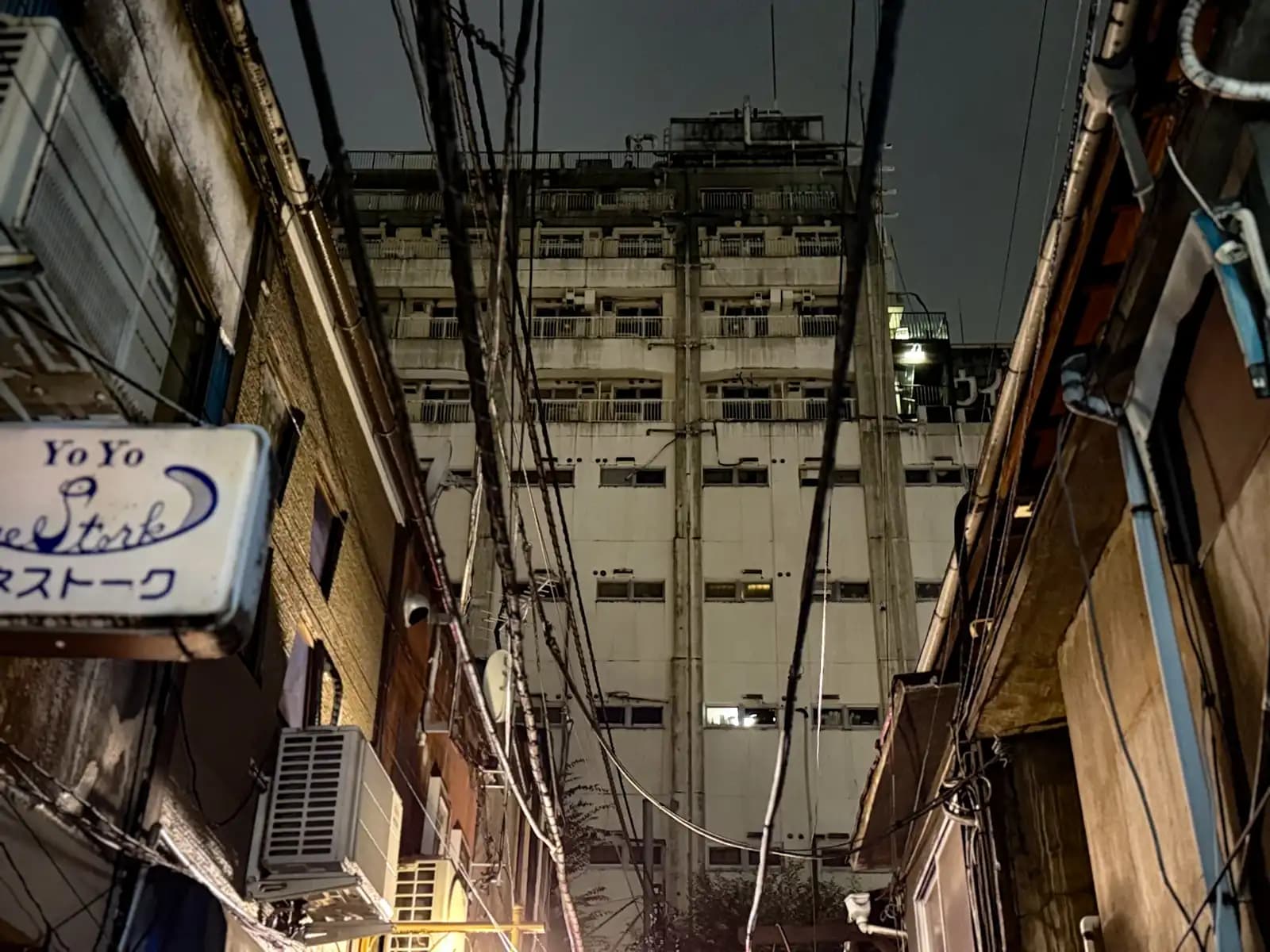 Looking up past old two-story bars and a sign for YoYo at a big concrete building towering over Golden Gai.