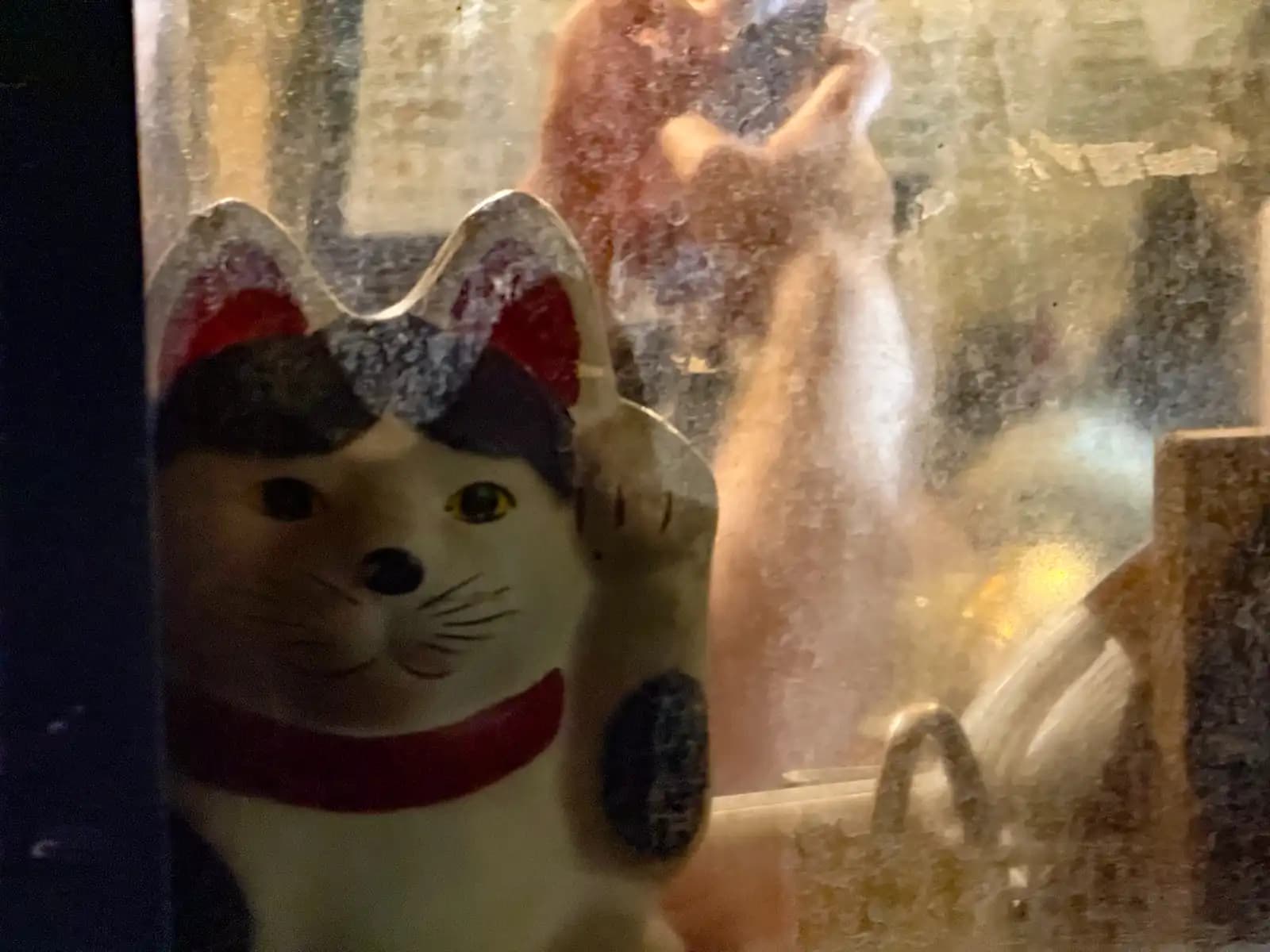 A maneki-neko lucky cat in a bar window, with someone inside checking their phone behind the dusty glass.
