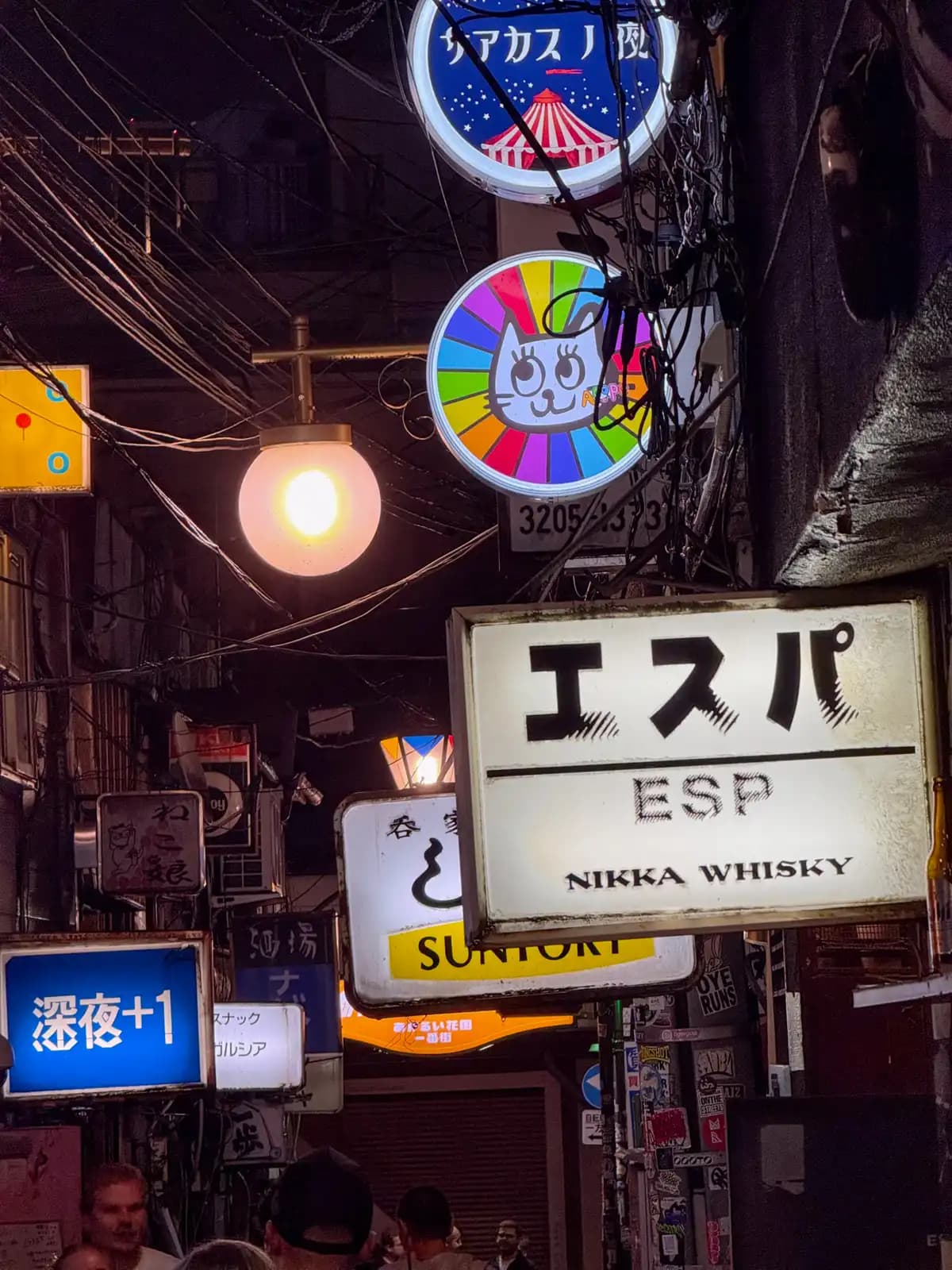 A stack of bar signs over a crowded alley, including Espa with a Nikka Whisky logo, a rainbow cat, and Shinya +1.