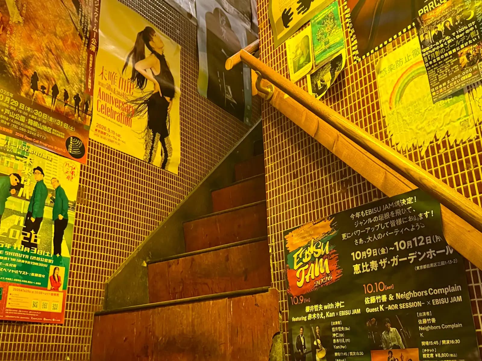 A narrow wooden staircase with a tiled wall full of concert posters, including one for Ebisu Jam 2026.