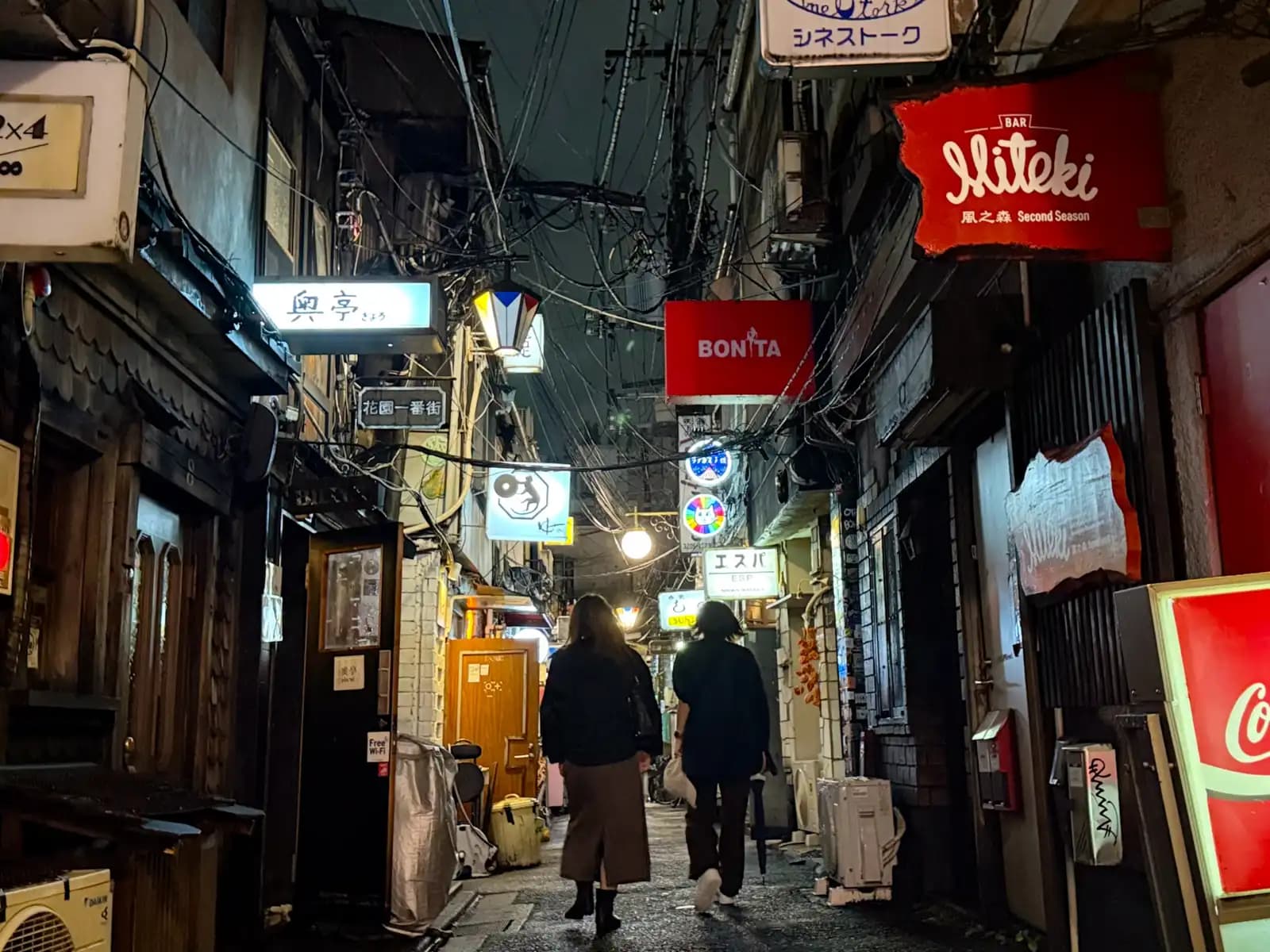 Two women walking down a wet alley past signs for Bar Miteki, Bonita and Espa.