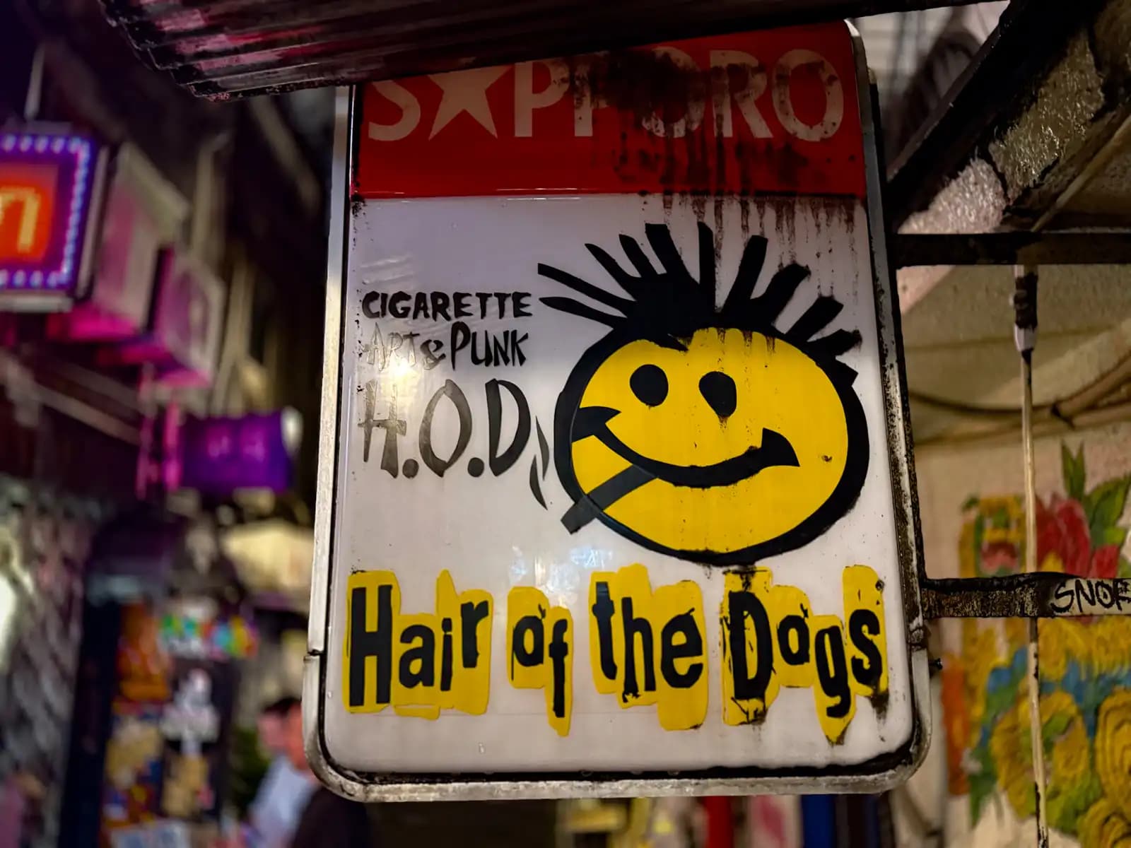 The Sapporo beer sign for Hair of the Dogs, a punk bar, with a spiky-haired smiley face smoking a cigarette.